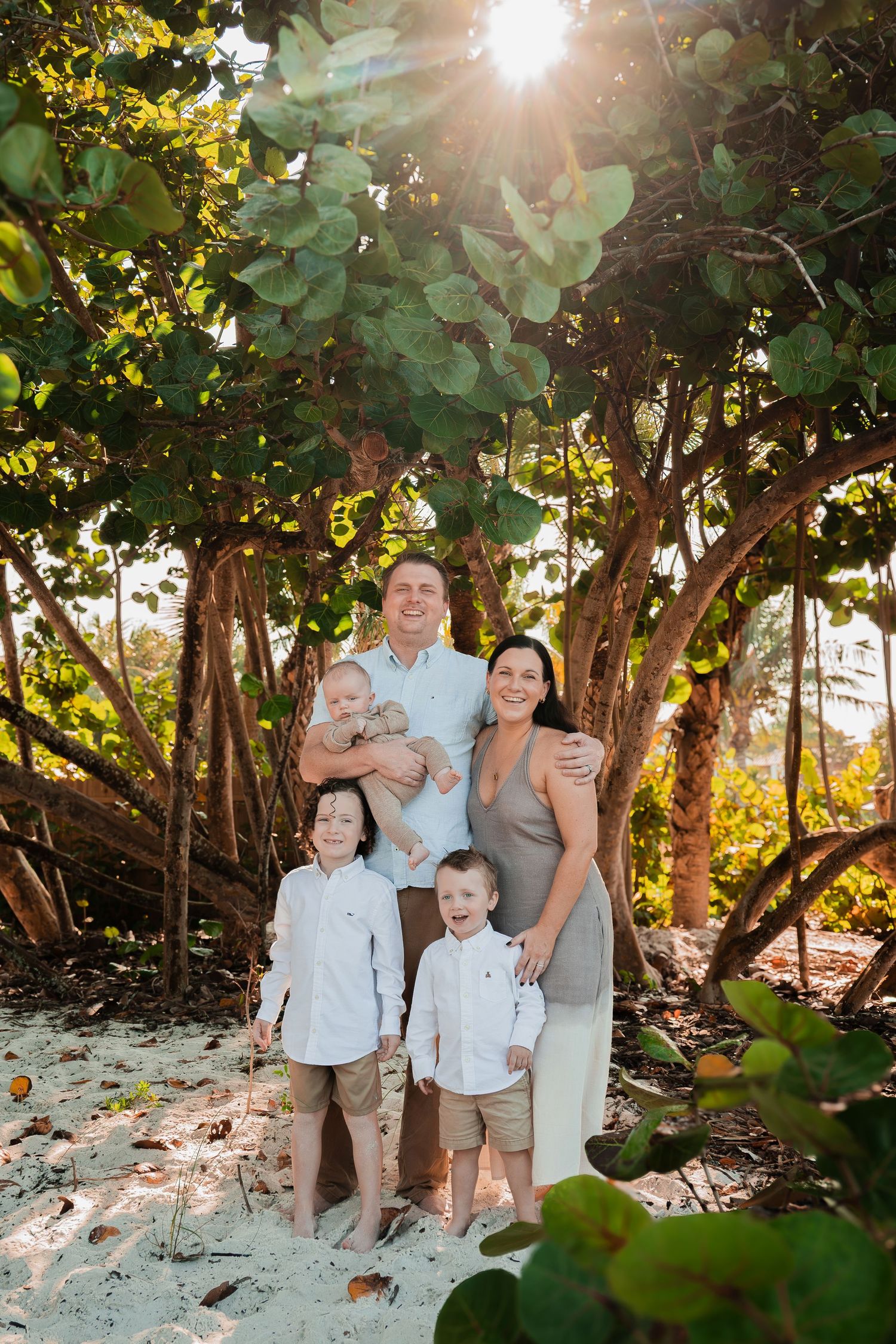 Family of five posing under lush green trees on a sunny beach, parents holding baby with two boys in white shirts.