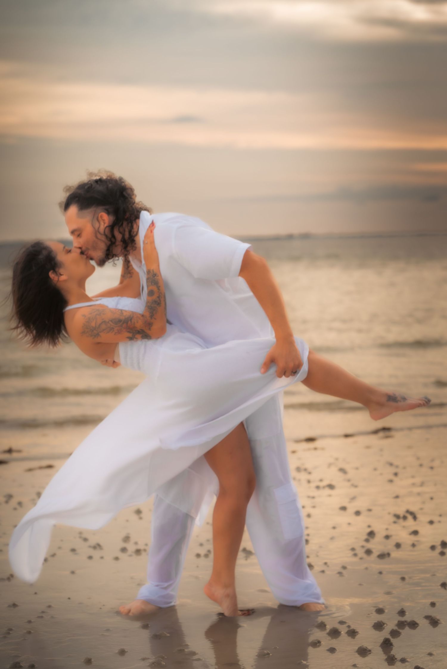 Couple sharing a romantic dip kiss on the beach at sunset, both wearing white outfits with tattoos visible.