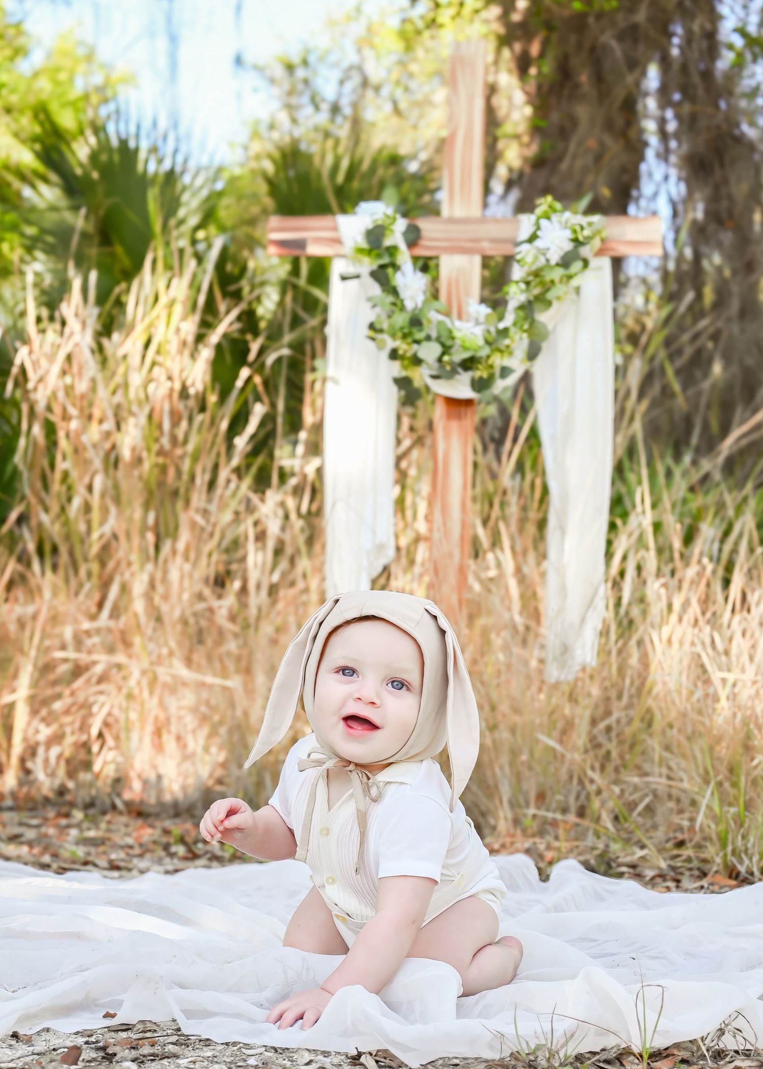 Cute baby in bunny bonnet sitting on white blanket with a wooden cross and greenery in background.