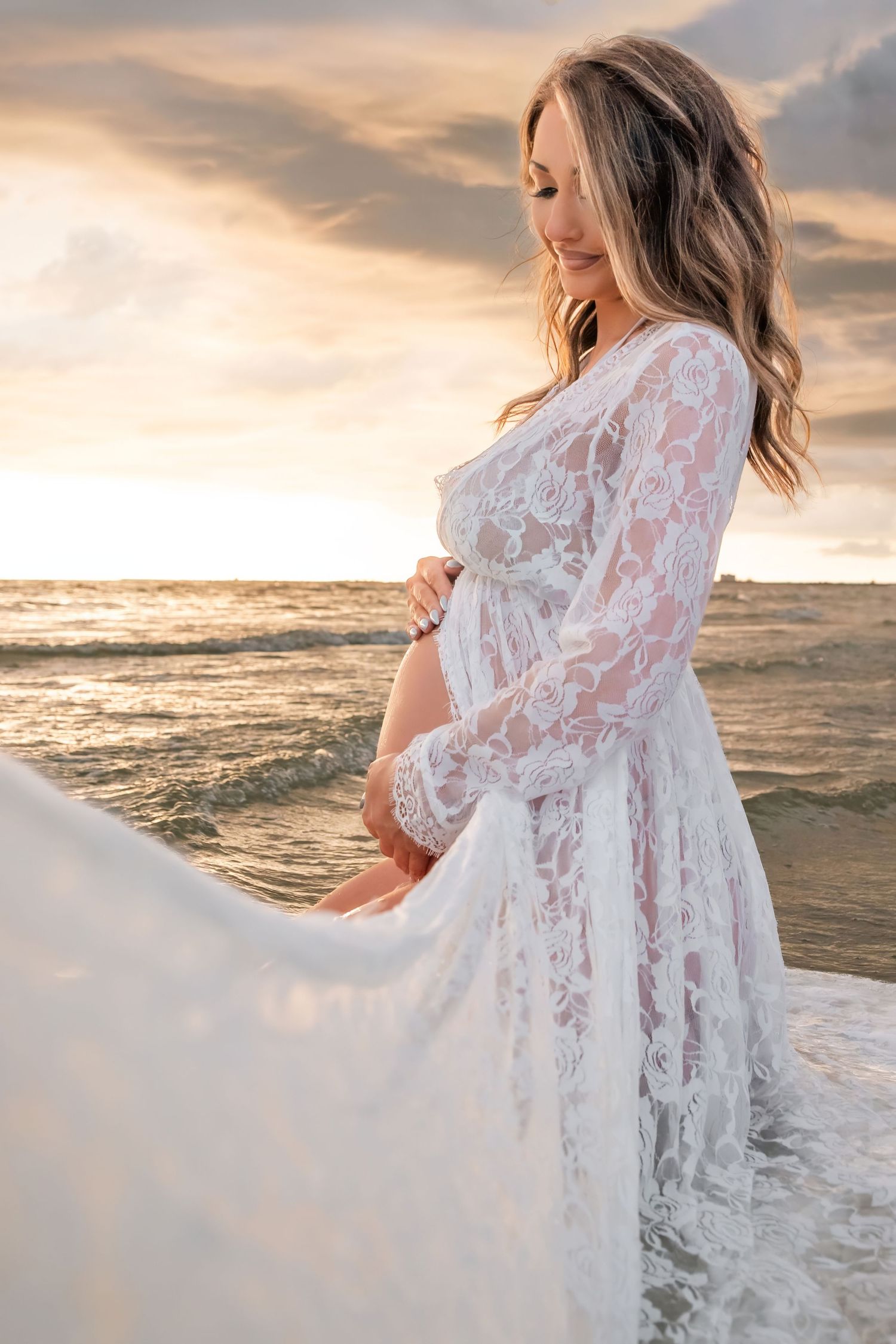 Pregnant woman in white lace dress standing on beach at sunset, cradling baby bump by ocean waves.