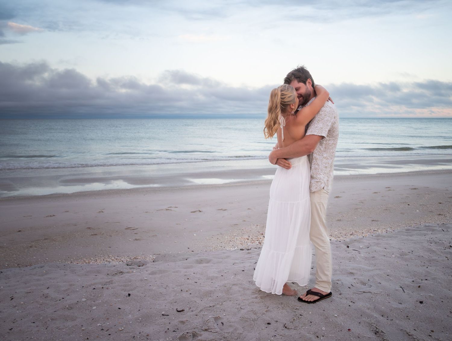 A couple in wedding attire embracing on a sandy beach at sunset with calm ocean waves in the background.