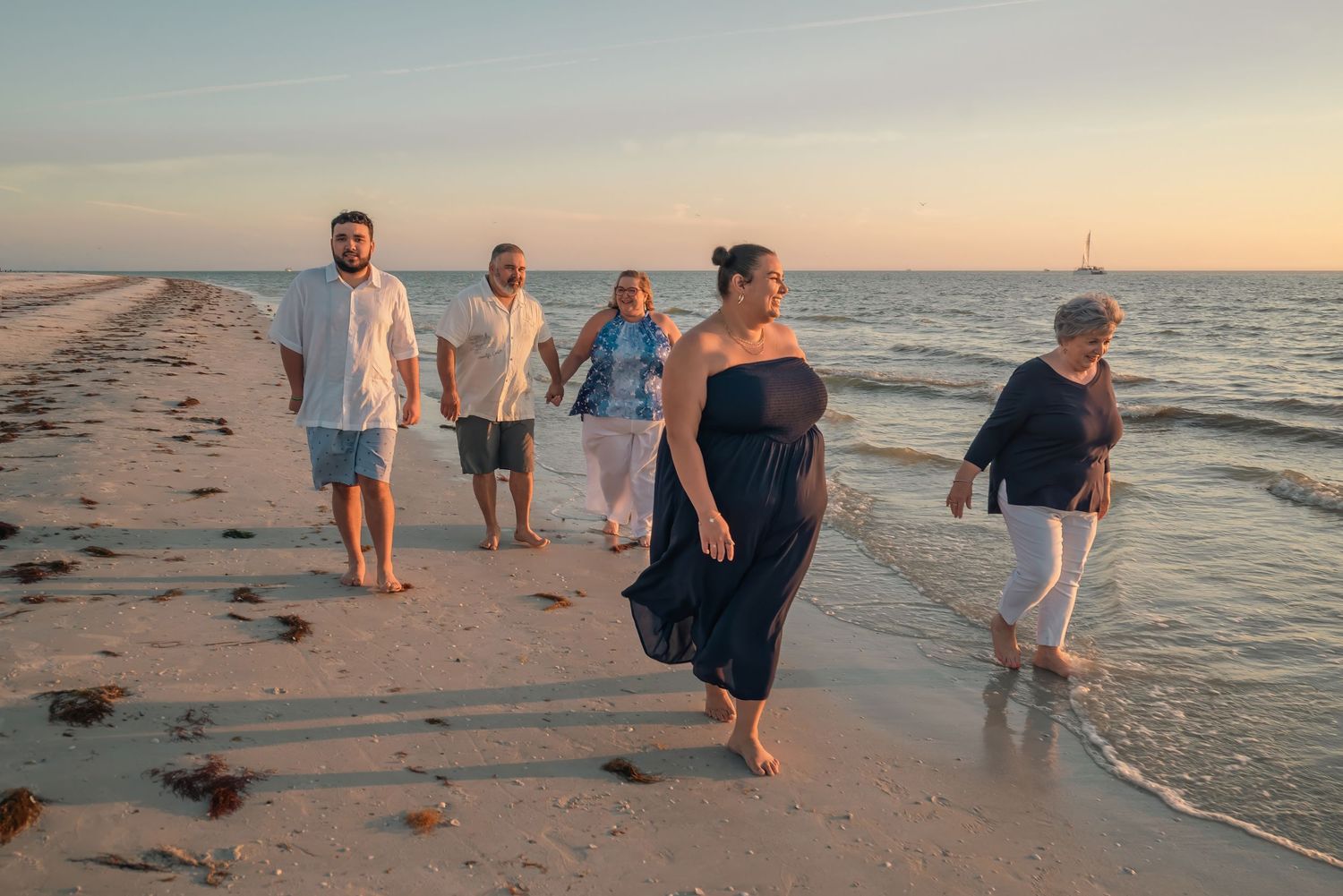 Family group of five casually walking along sandy beach shoreline during a colorful sunset.