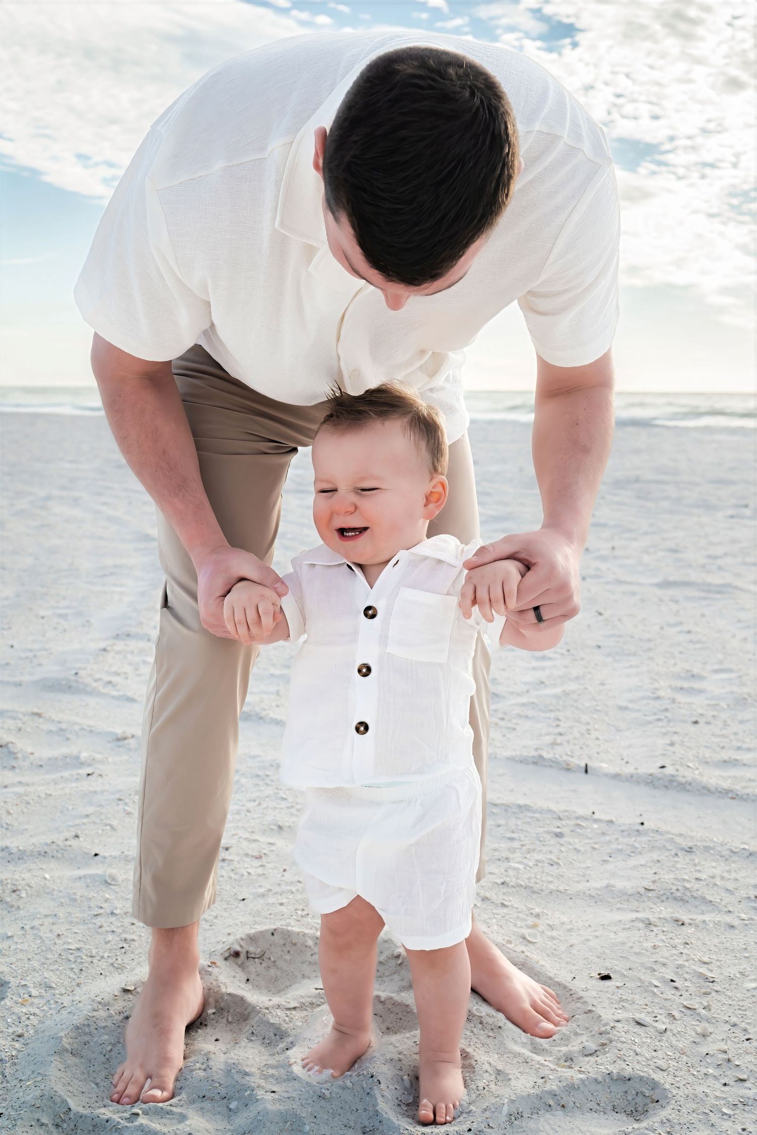 A father helps his laughing baby boy stand on sandy beach under cloudy skies.