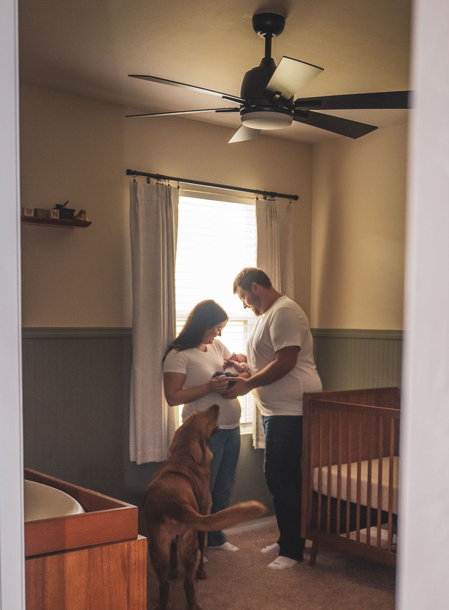 New parents holding newborn in nursery as family dog looks on beside a wooden crib.