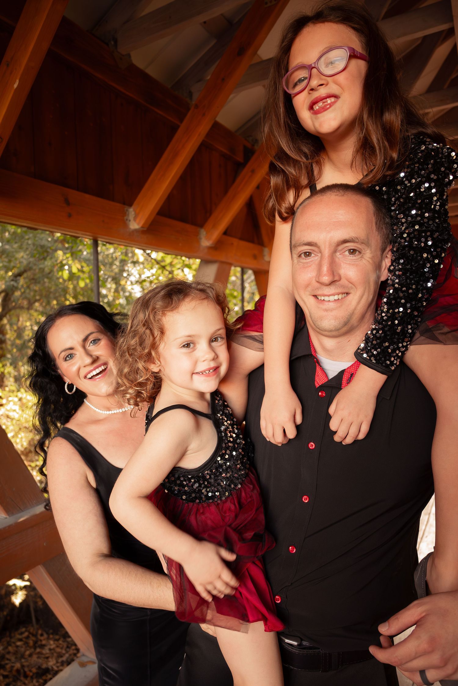 Happy family of four poses under a wooden pavilion with two daughters in festive black and red outfits.