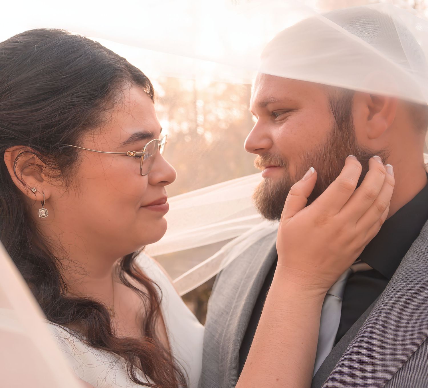 Bride and groom share an intimate moment under wedding veil in soft sunlight.