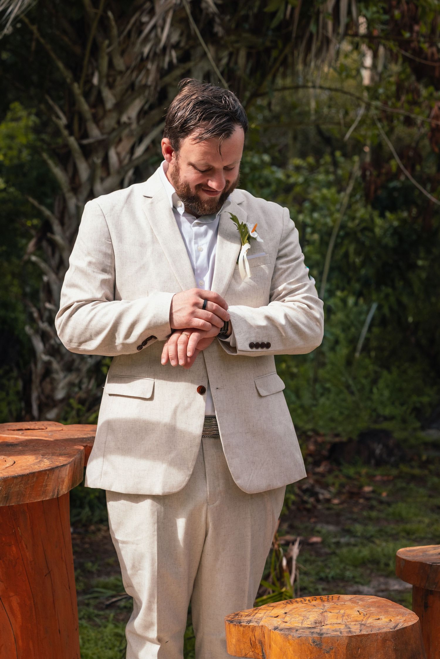 Groom in beige linen suit adjusting cufflinks outdoors, wearing boutonniere, with wooden stumps and greenery background.
