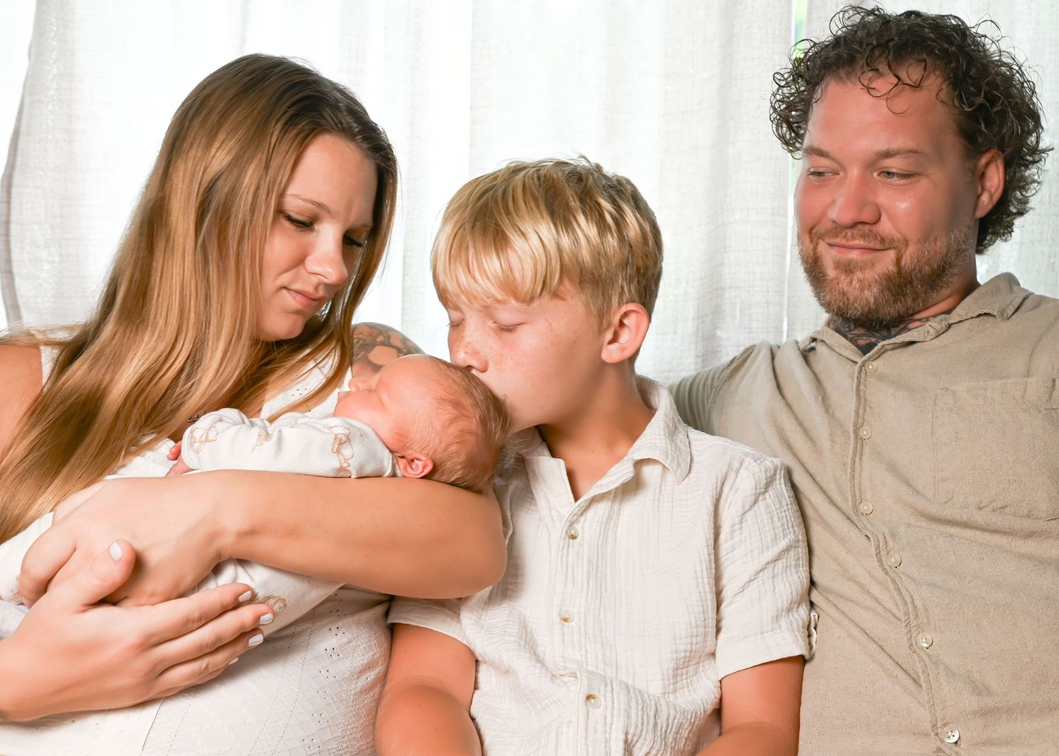 Family portrait with mother holding newborn baby, young boy kissing infant, and father smiling warmly together.