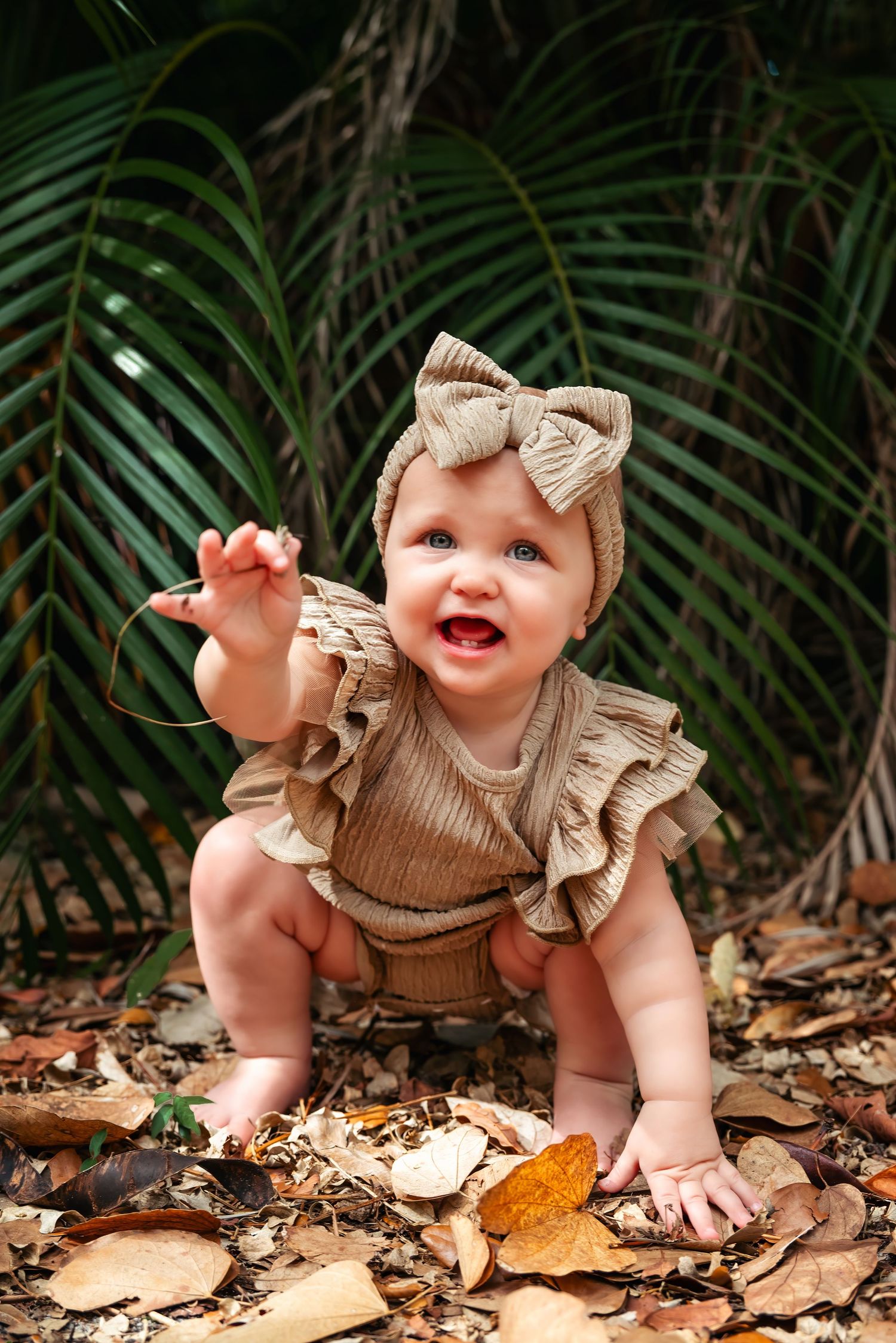 Adorable baby girl in tan ruffle romper and bow headband sitting among tropical leaves outdoors.