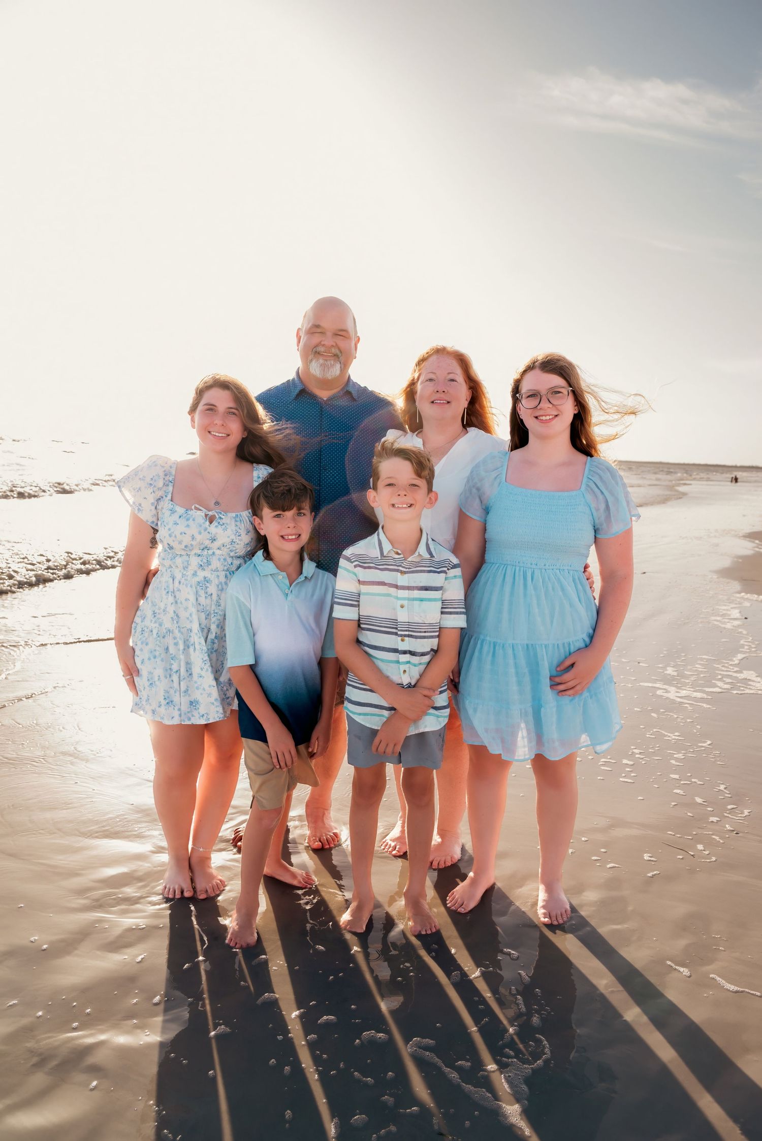Family of six posing together on a sandy beach at sunset, wearing coordinating blue and white outfits.