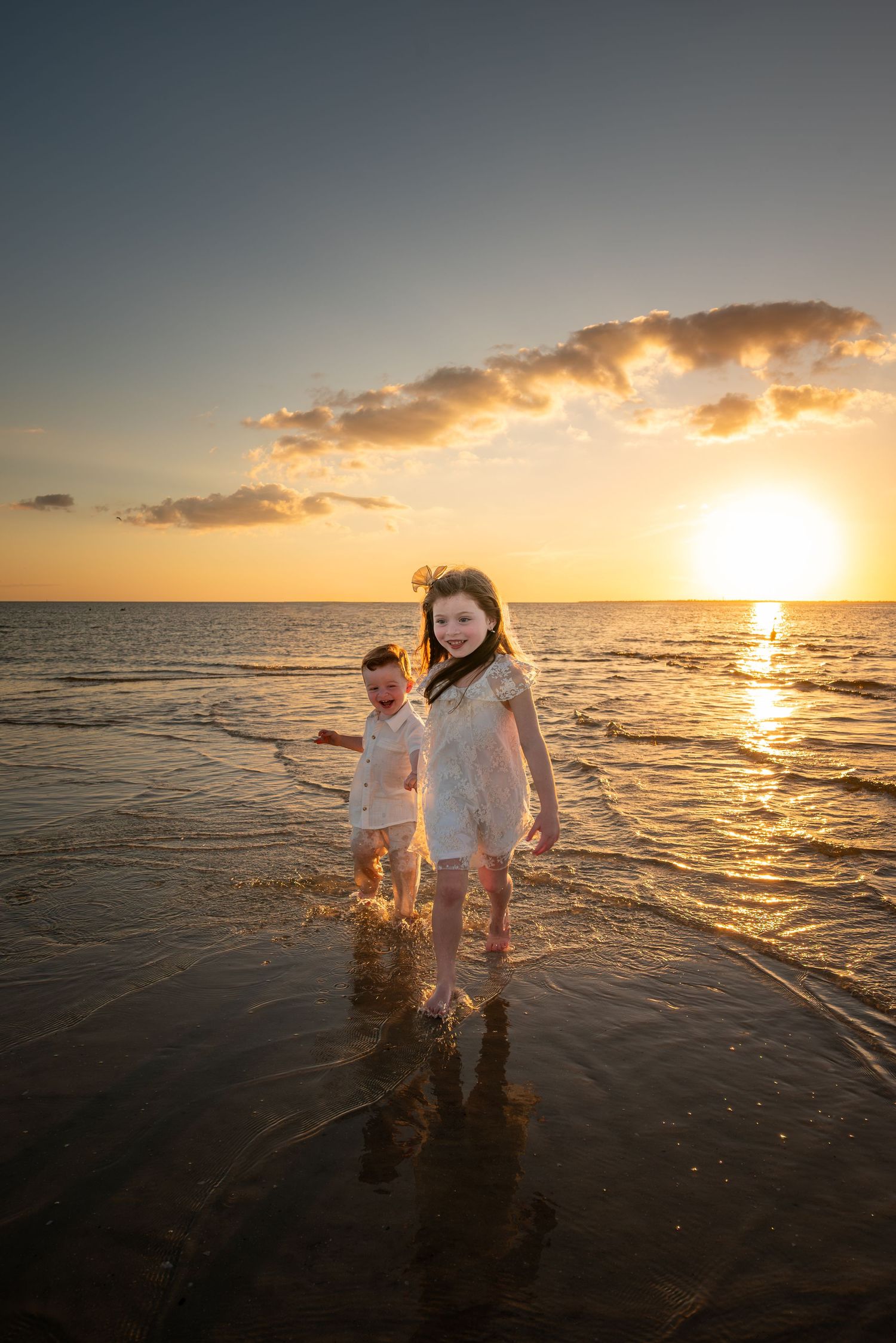 Two children in white clothes play in ocean waves during a stunning golden sunset at the beach.