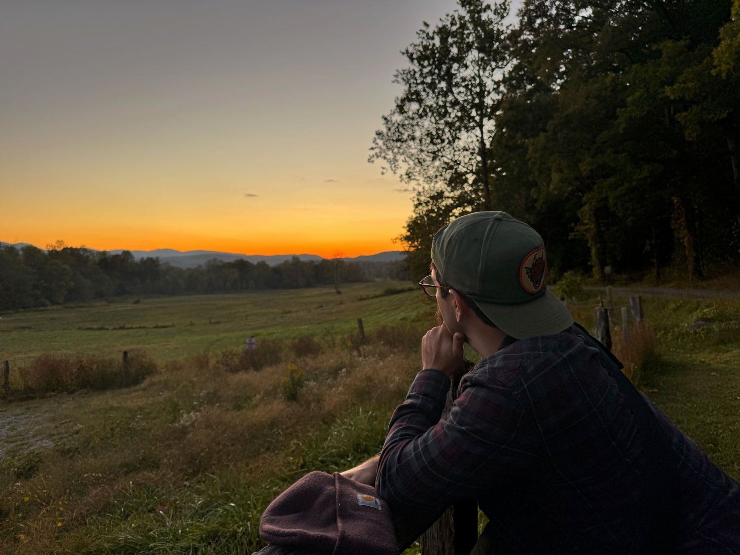 Person in green cap watches vibrant orange sunset over rural valley landscape with trees and fields.