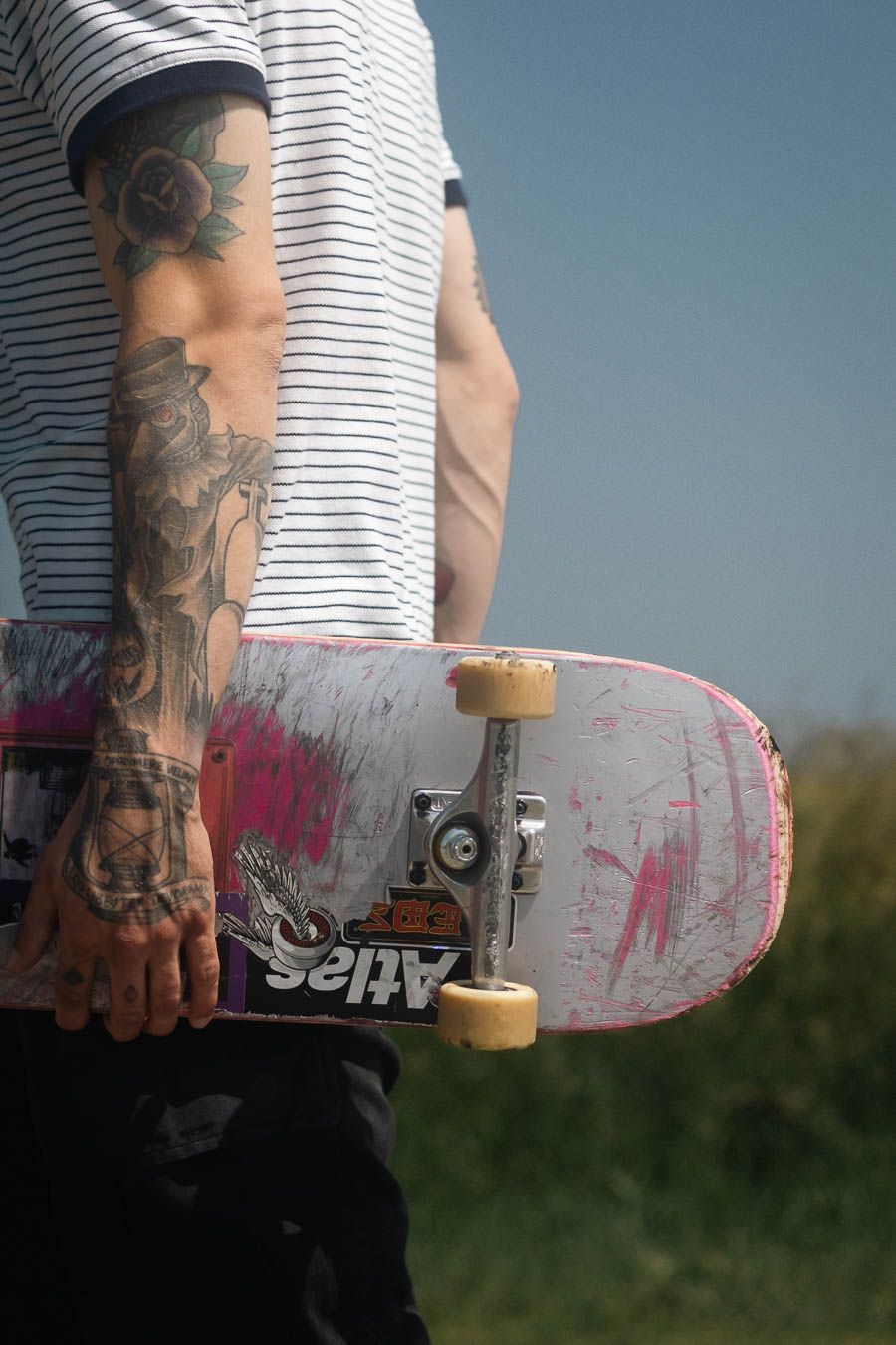 Close-up view of a tattooed arm holding a worn pink skateboard deck against a grassy outdoor background.