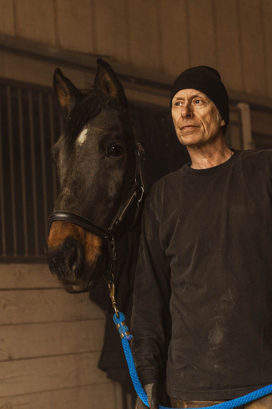 A person in dark clothing stands next to a black horse with a blue lead rope in a dimly lit stable.
