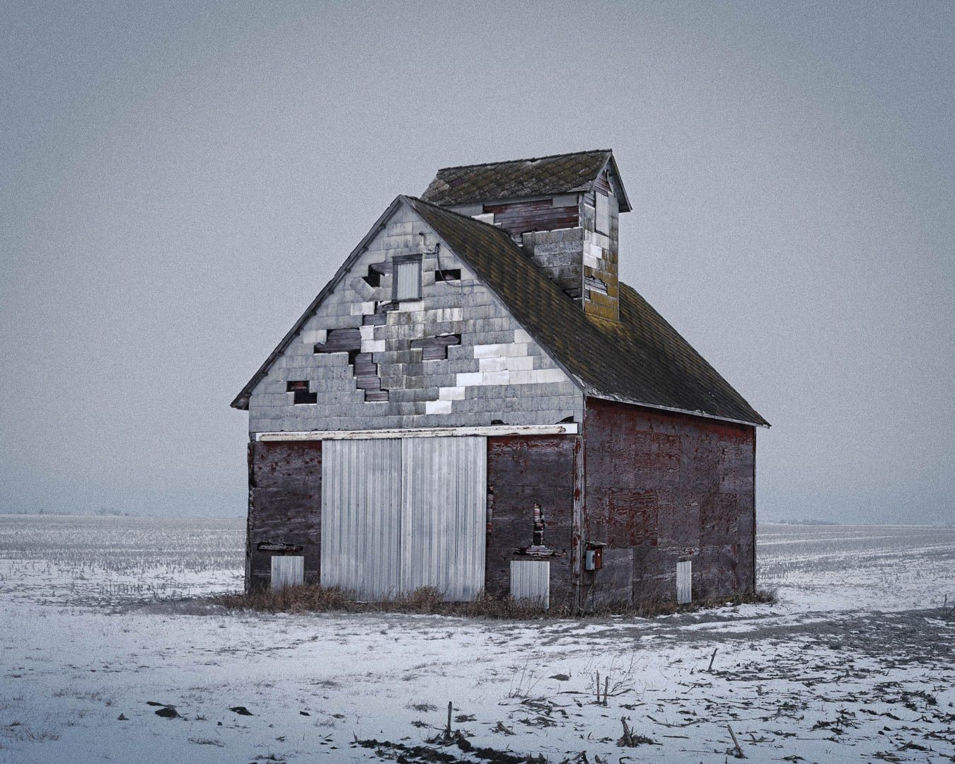 A series of abandoned wooden barns stand weathered and dilapidated in a snowy winter landscape.