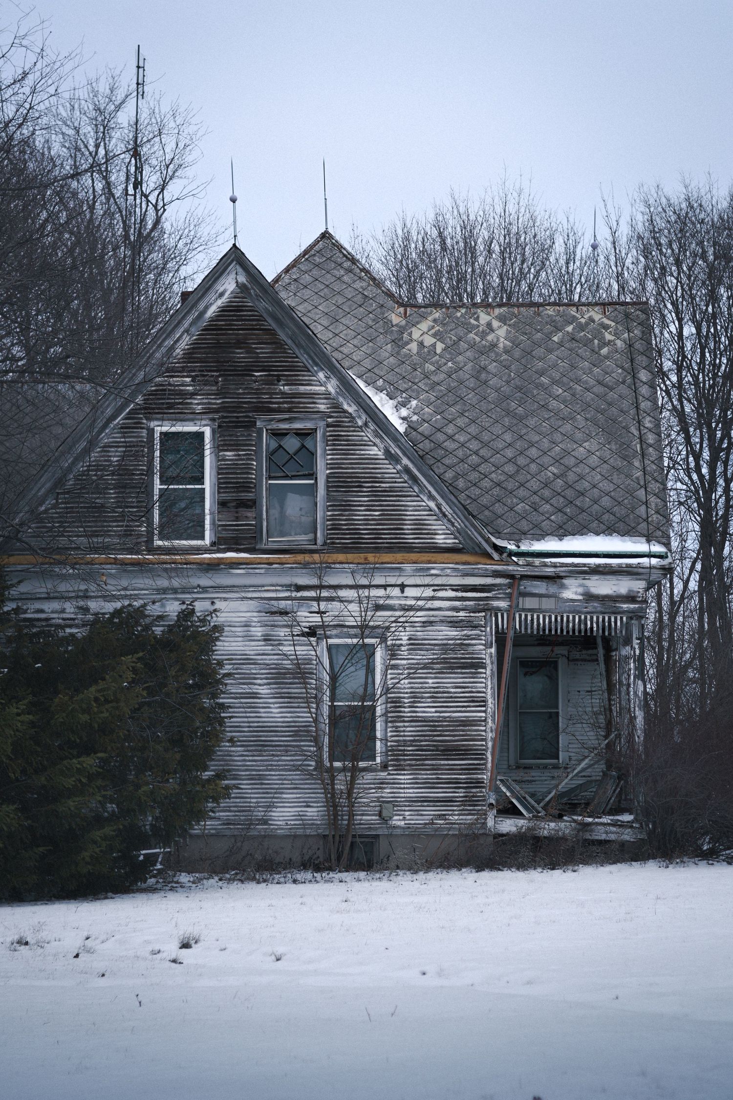 Abandoned farmhouse with weathered siding sits in snowy field surrounded by bare winter trees and grey sky.