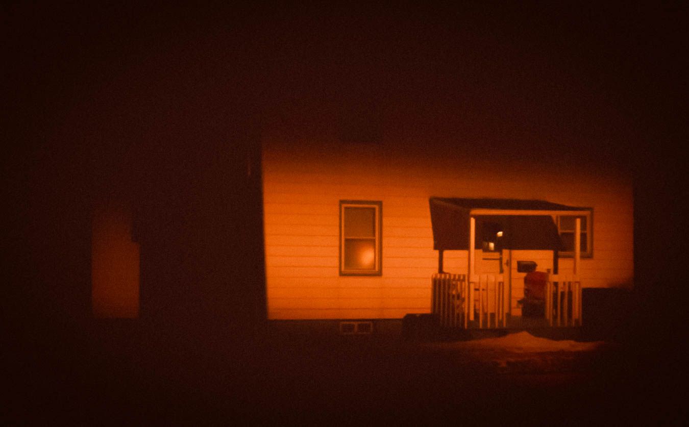 A dimly lit house exterior at night with an orange glow illuminating its wooden siding and front porch.