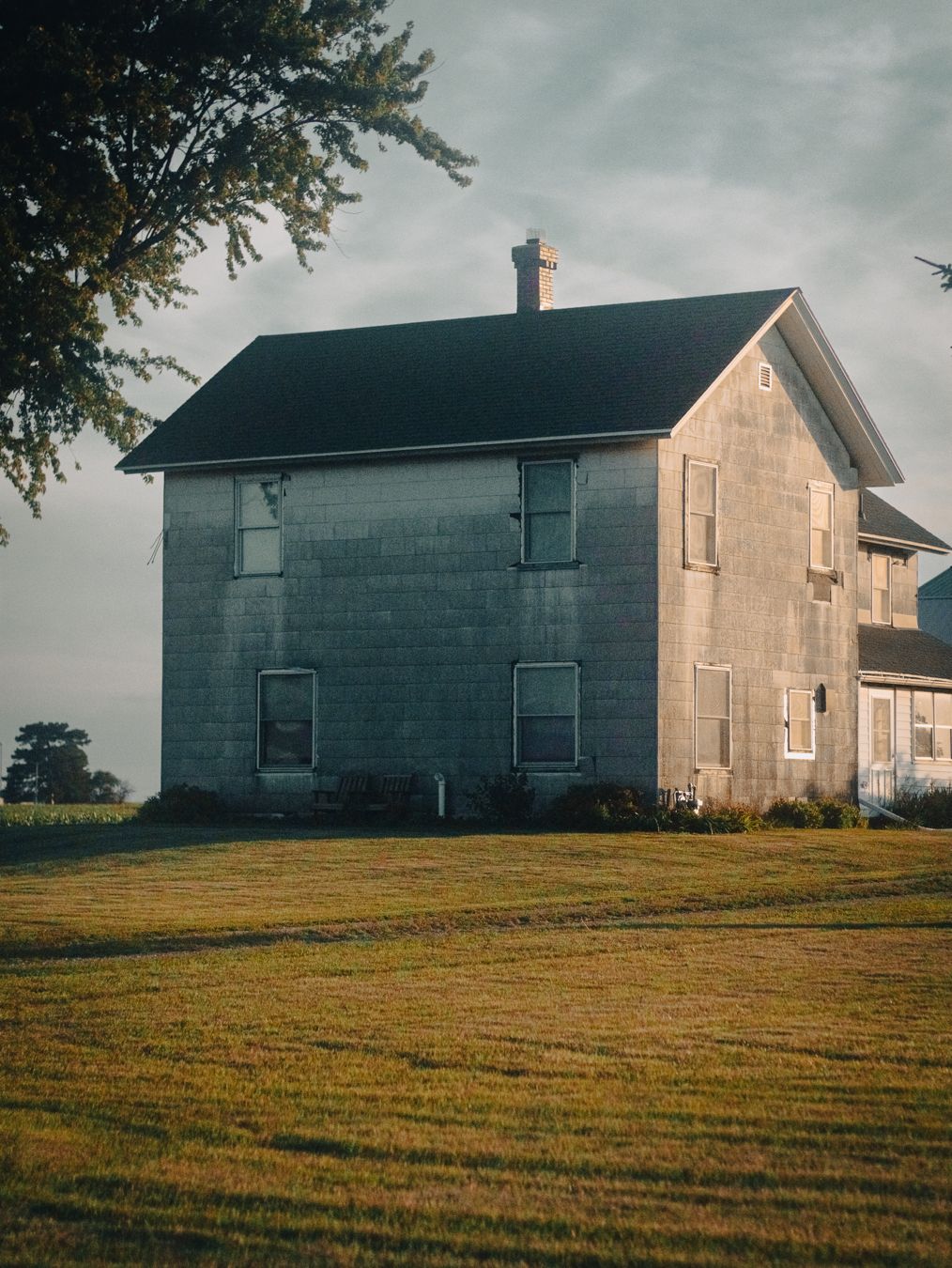 A rustic two-story farmhouse with gray siding and dark roof sits on a green lawn under a cloudy sky.