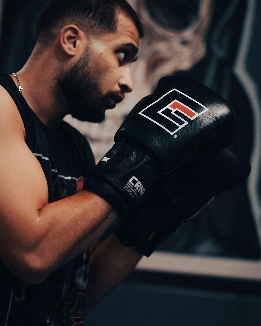 A male boxer in black boxing gloves practices fighting stance in a gym wearing a sleeveless shirt.