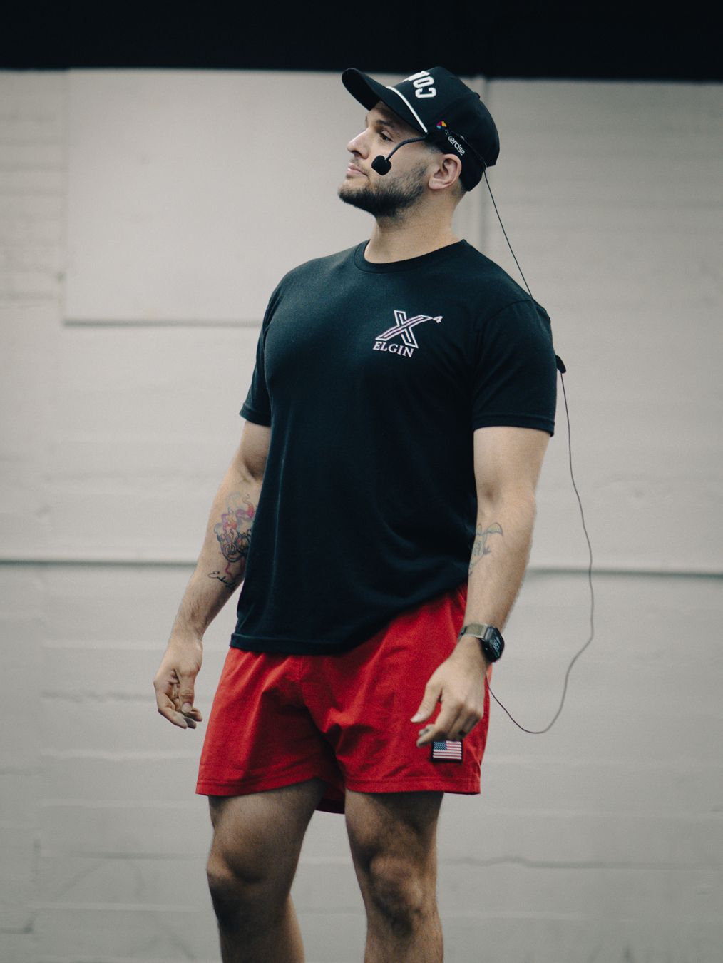 Muscular man wearing black t-shirt, red shorts, and black cap with headset microphone stands confidently indoors.
