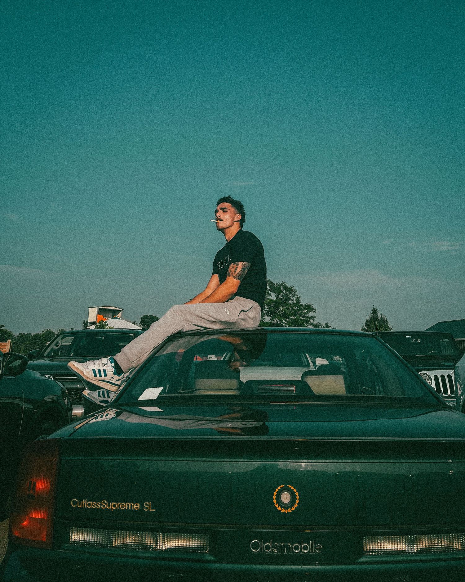Young man with tattoos sitting on top of a green Oldsmobile Cutlass Supreme SL in a parking lot at dusk.