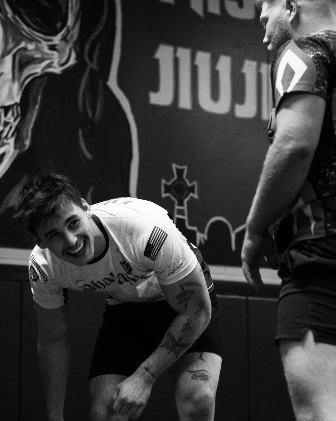Smiling tattooed man in white tee at a Jiu-Jitsu gym, black and white photo.