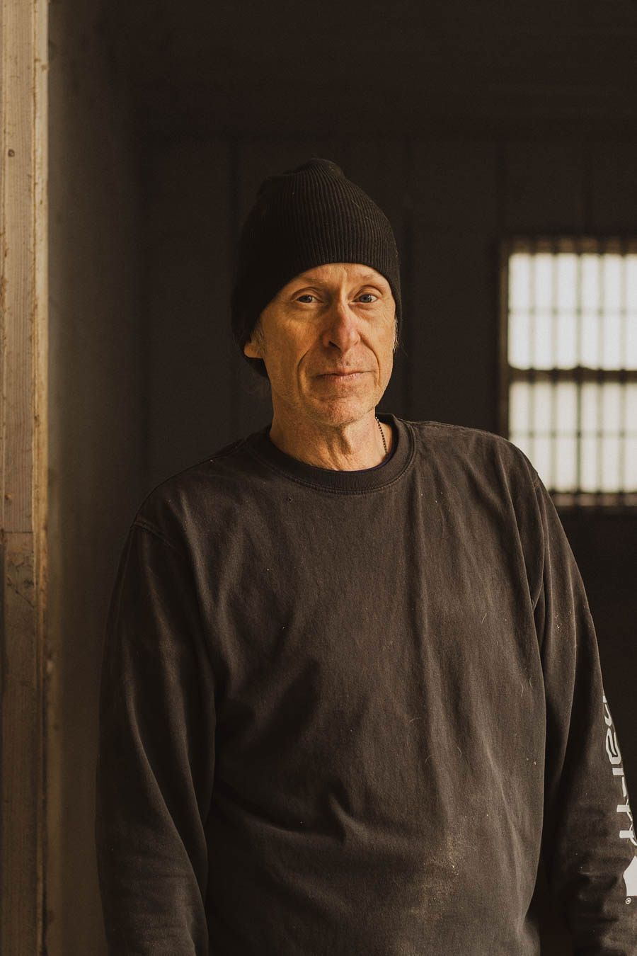 Person wearing dark winter clothing and beanie smiles while standing in dimly lit room with window in background.