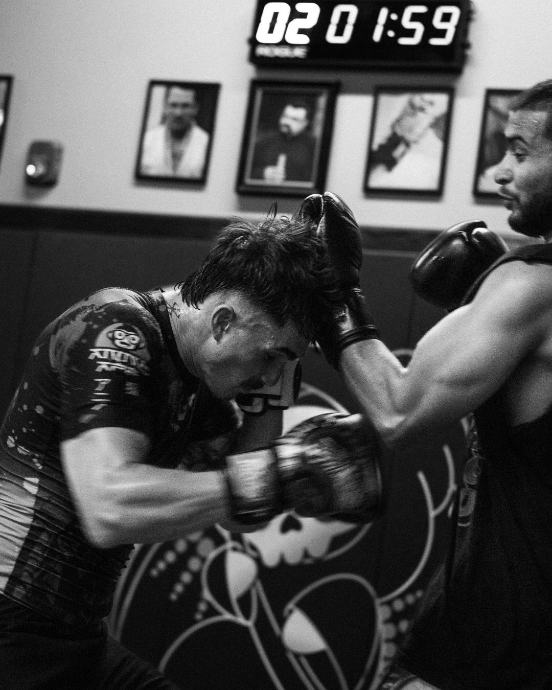 Two MMA fighters sparring in a gym with a countdown timer showing 1:59 on the wall in a black and white photo.