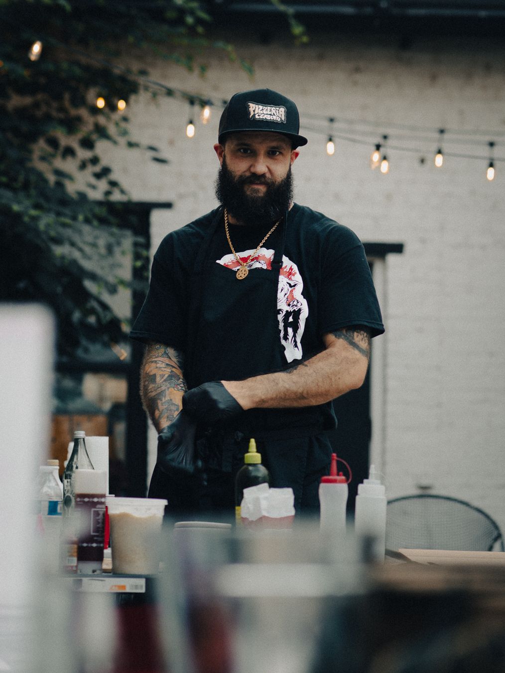 Tattooed male chef in black cap and gloves prepares food at outdoor station with string lights backdrop.