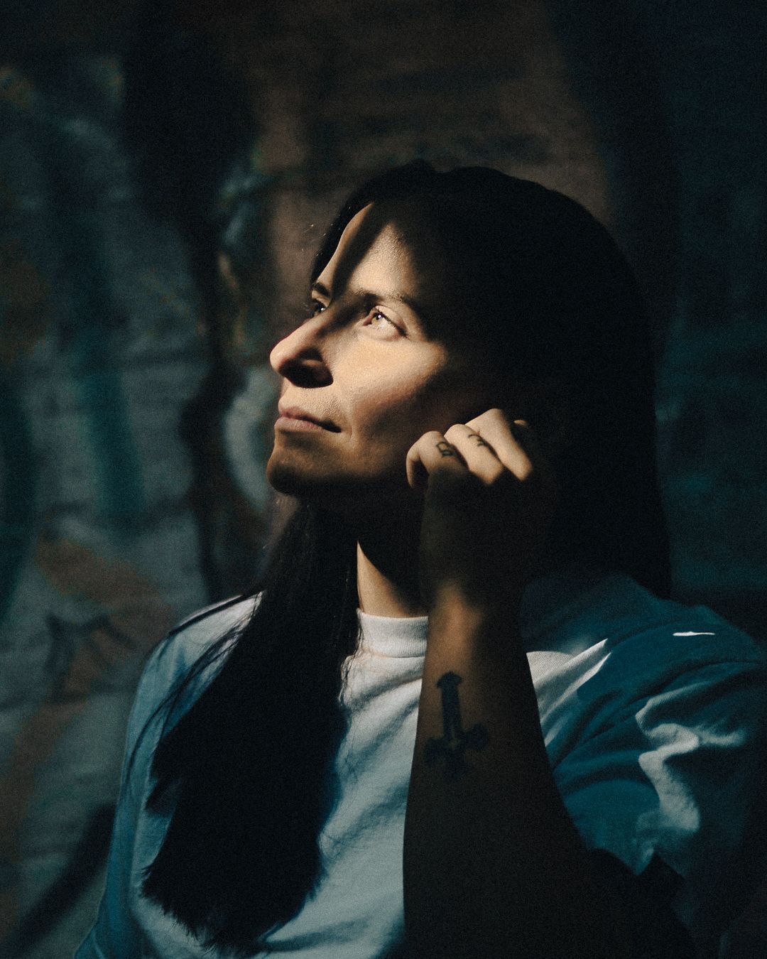Person with long dark hair and tattoo on forearm gazes upward thoughtfully in dramatic moody lighting.