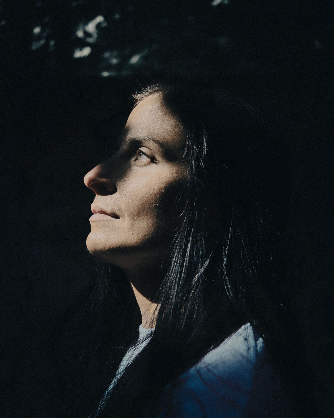 A woman with dark hair gazes upward in dramatic side lighting against a dark background.