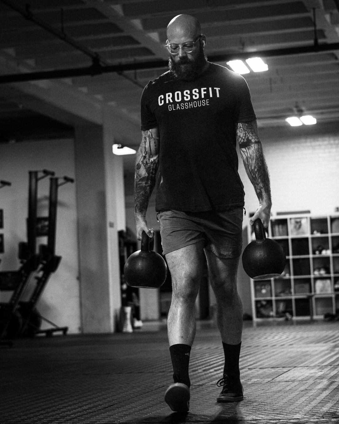 Bald tattooed man carries two kettlebells in a CrossFit Glasshouse gym, black and white photo.