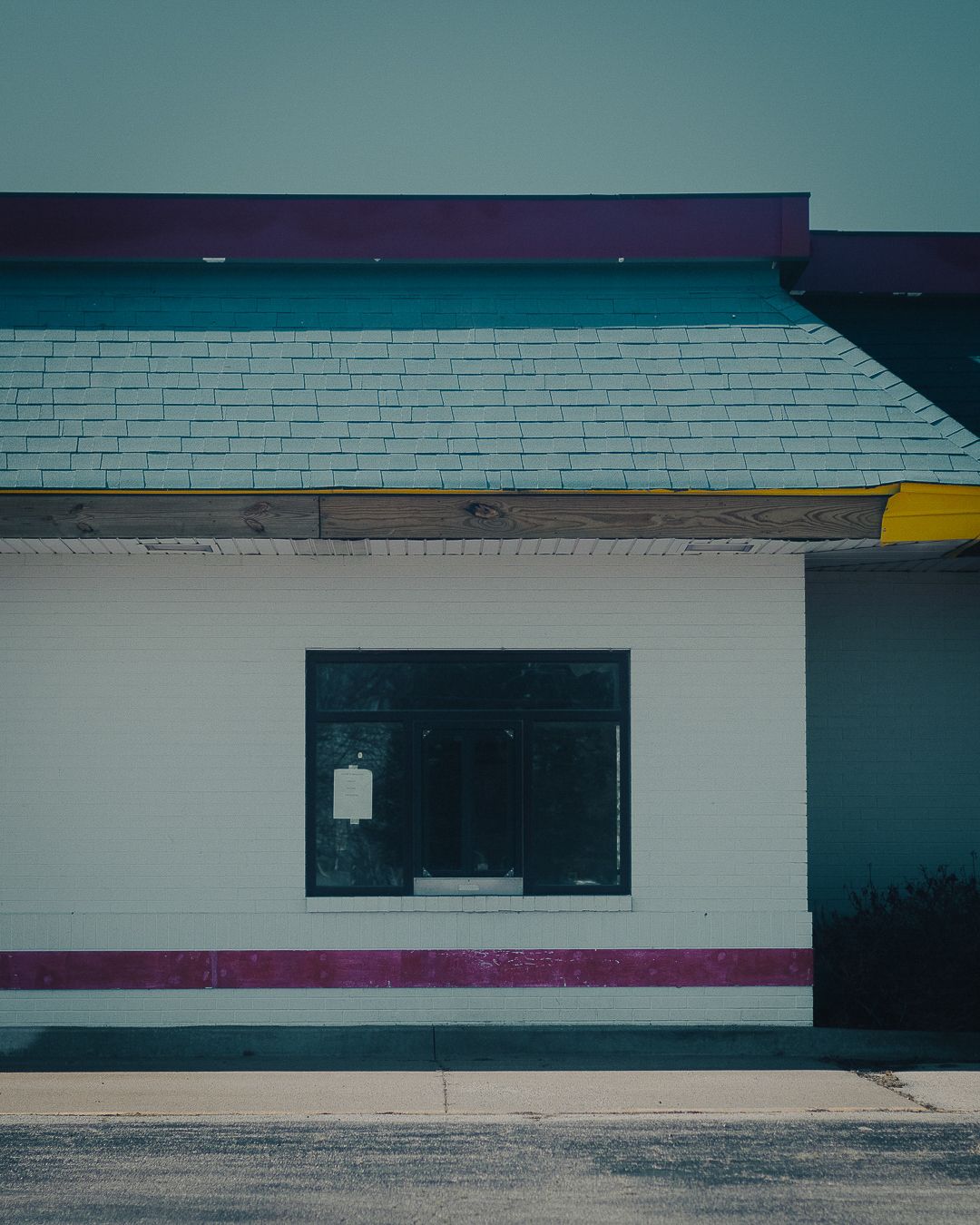 Abandoned fast food restaurant exterior with teal roof, white brick walls, and magenta stripe under large dark window.