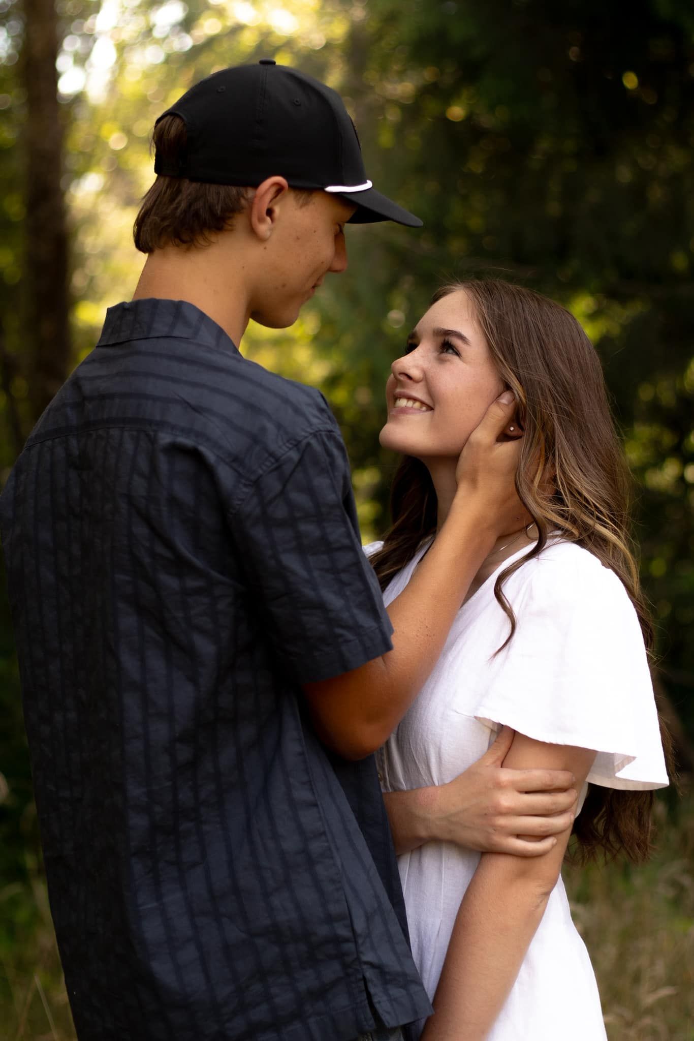 A romantic outdoor moment between two young people as they embrace in a sunlit wooded setting.