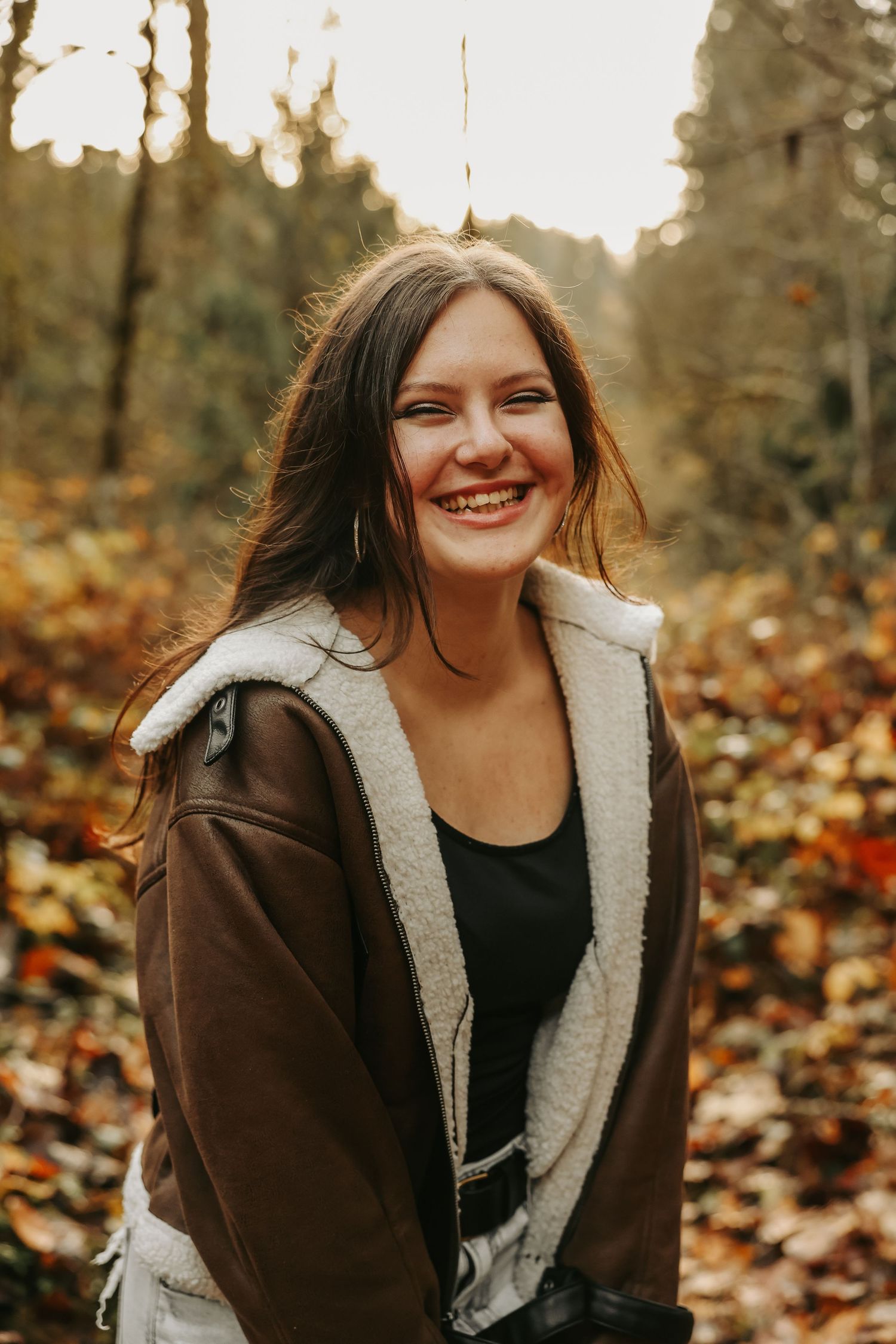 A person in a brown sherpa jacket smiles warmly while sitting among autumn leaves in a wooded setting.