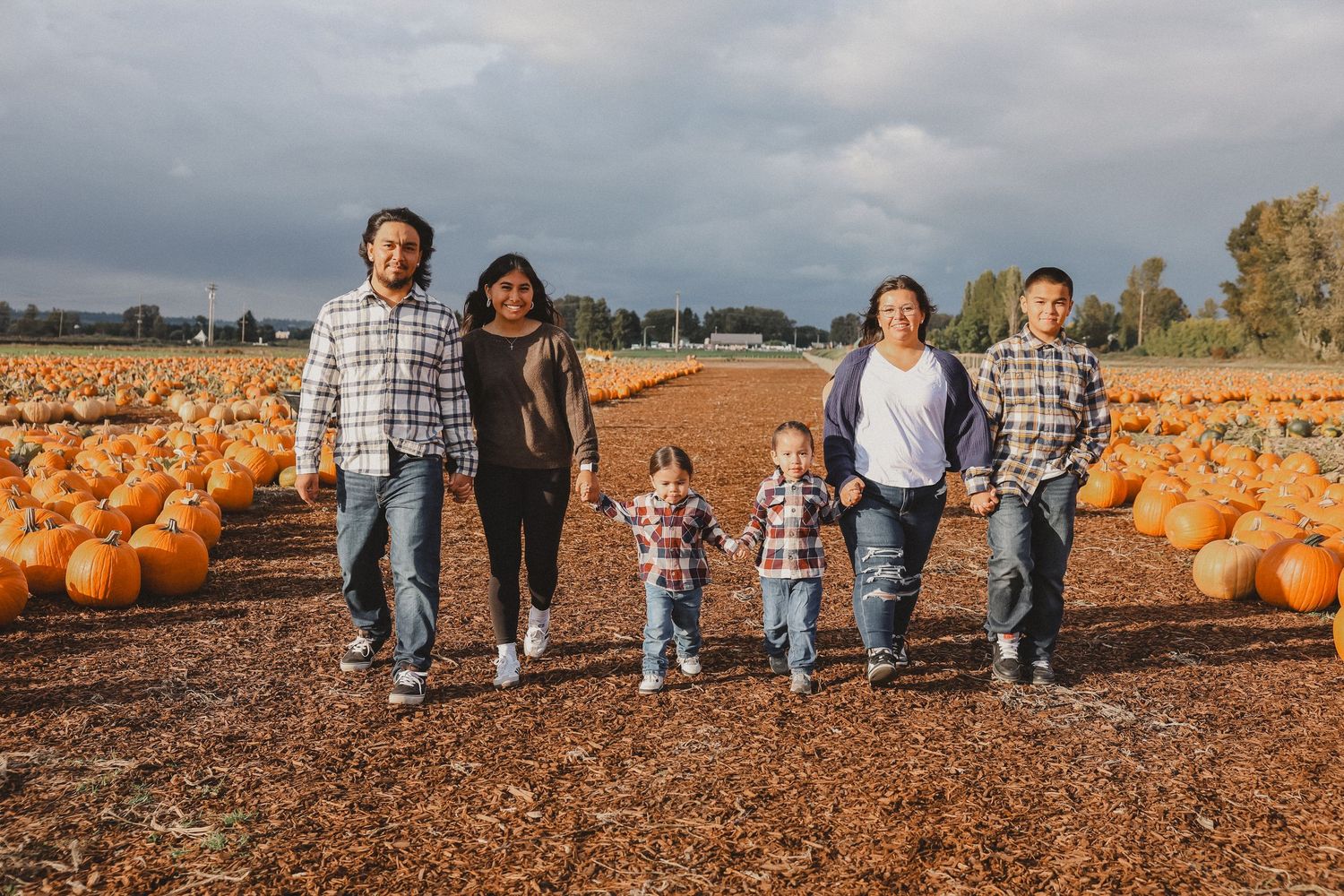 A family walks together through a pumpkin patch filled with orange pumpkins on a cloudy autumn day.