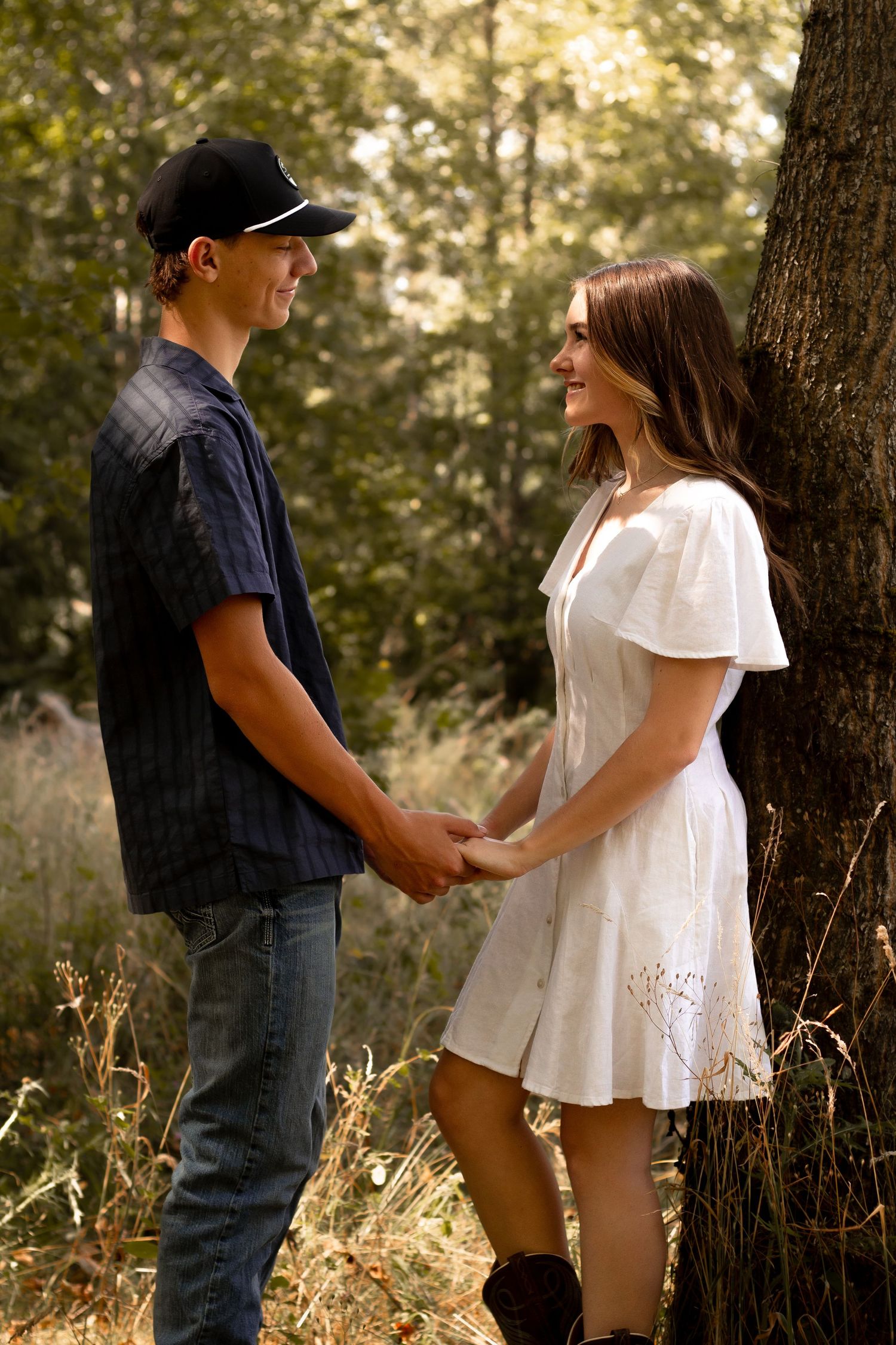 A couple holds hands while standing in a sunlit forest setting with golden light filtering through the trees.