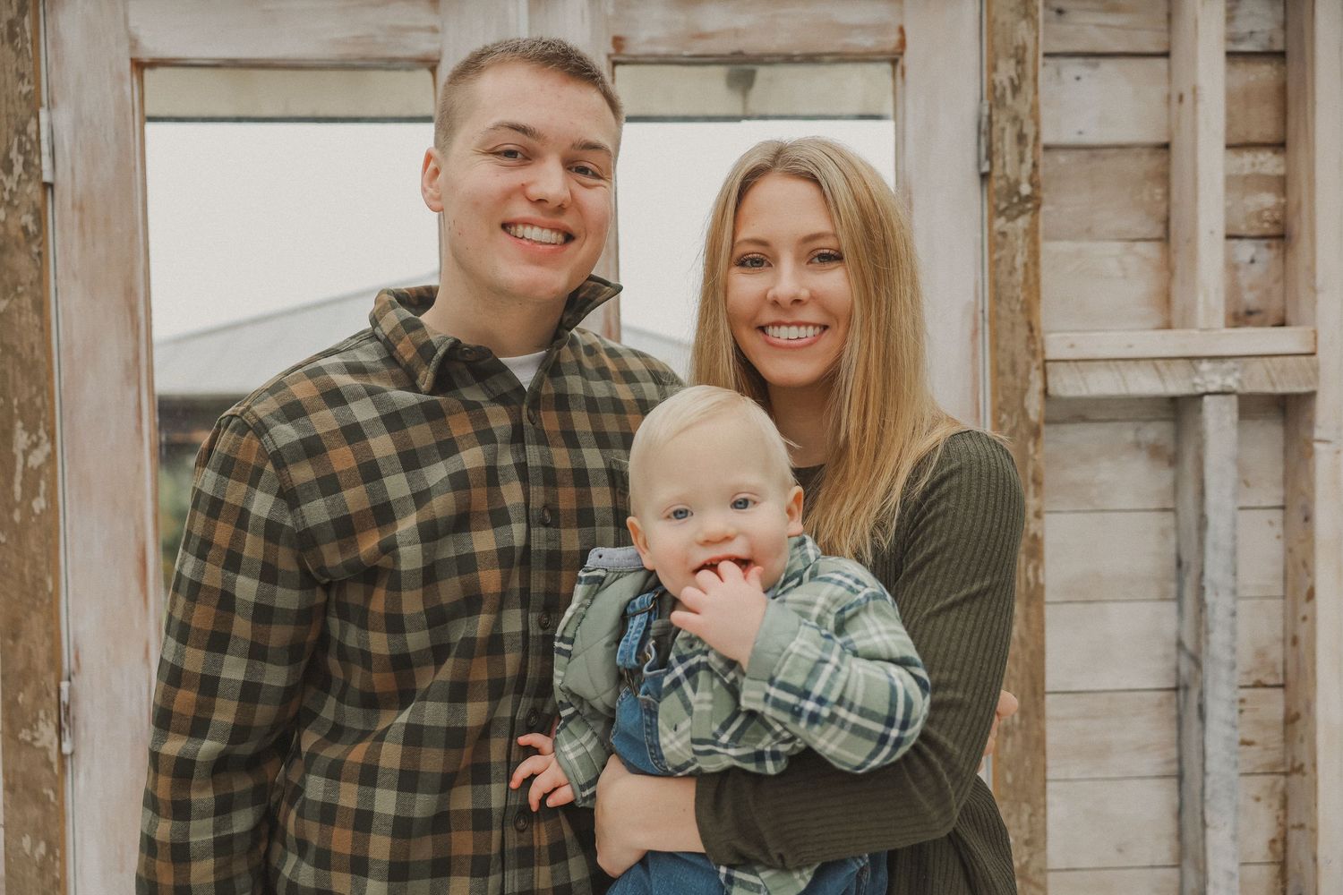 Family poses together in coordinated outfits against rustic wooden wall background during outdoor photo session.
