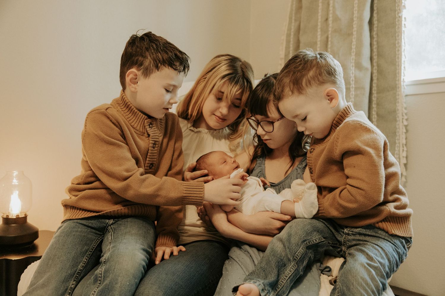 A mother sits with her three children, two boys and a girl, tenderly cradling a newborn baby in a warm, cozy setting.