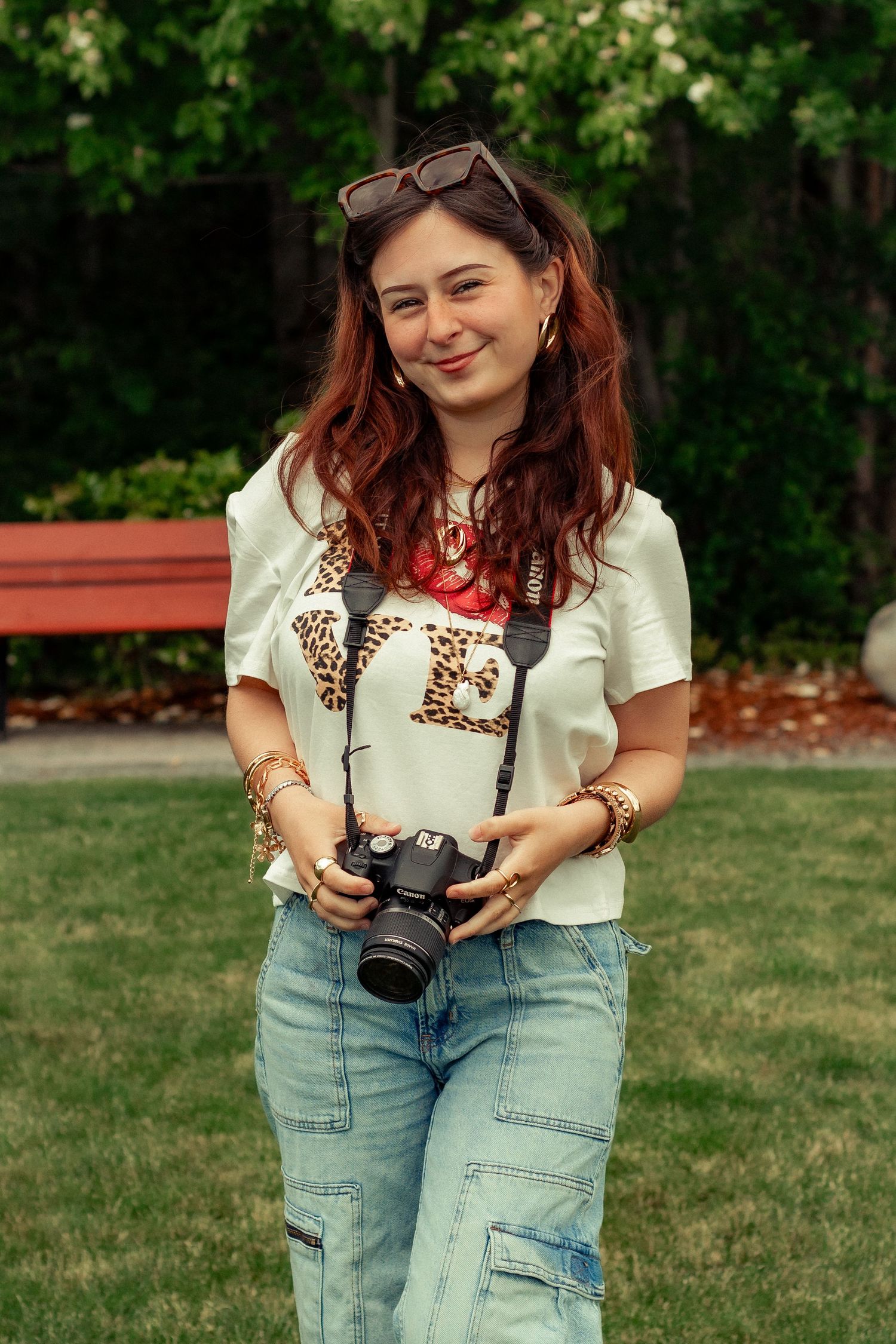Photographer holds a camera while wearing casual attire with sunglasses on head in an outdoor park setting.