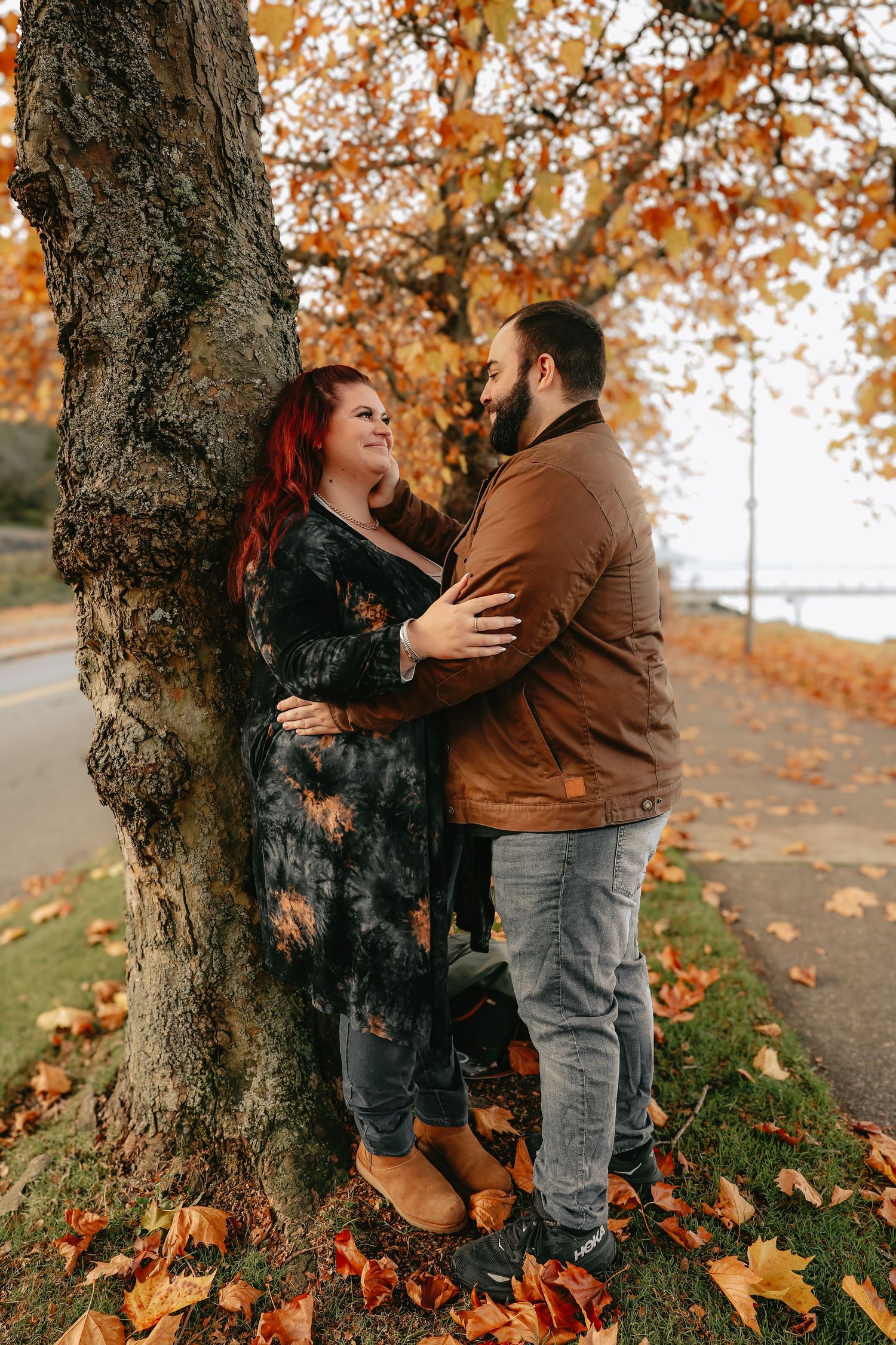 A couple embraces against a tree trunk surrounded by fallen autumn leaves on a peaceful path.