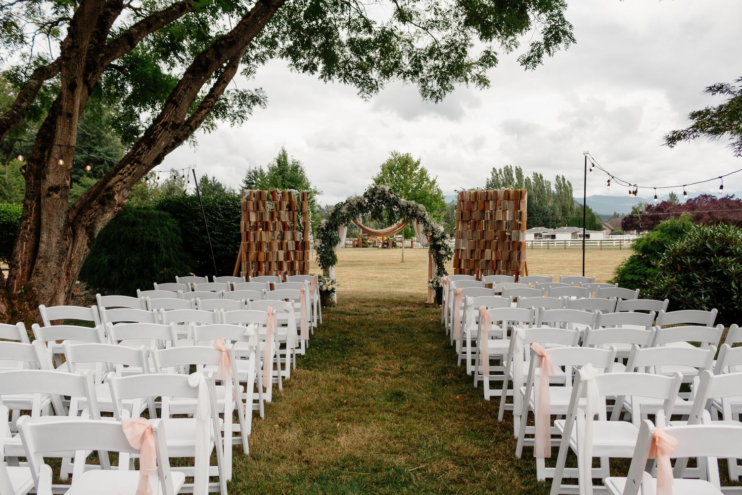 White wooden chairs arranged in rows for an outdoor wedding ceremony under tall trees and string lights.