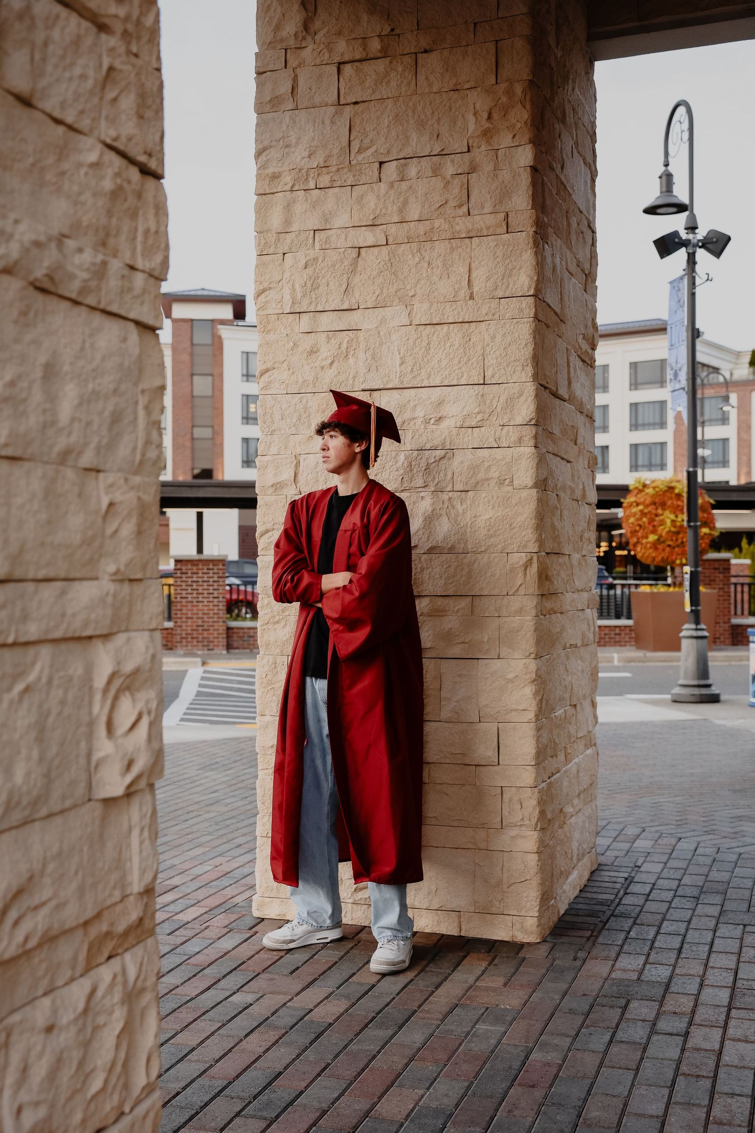 Graduate in red cap and gown stands against stone pillar on brick walkway with autumn trees in background.