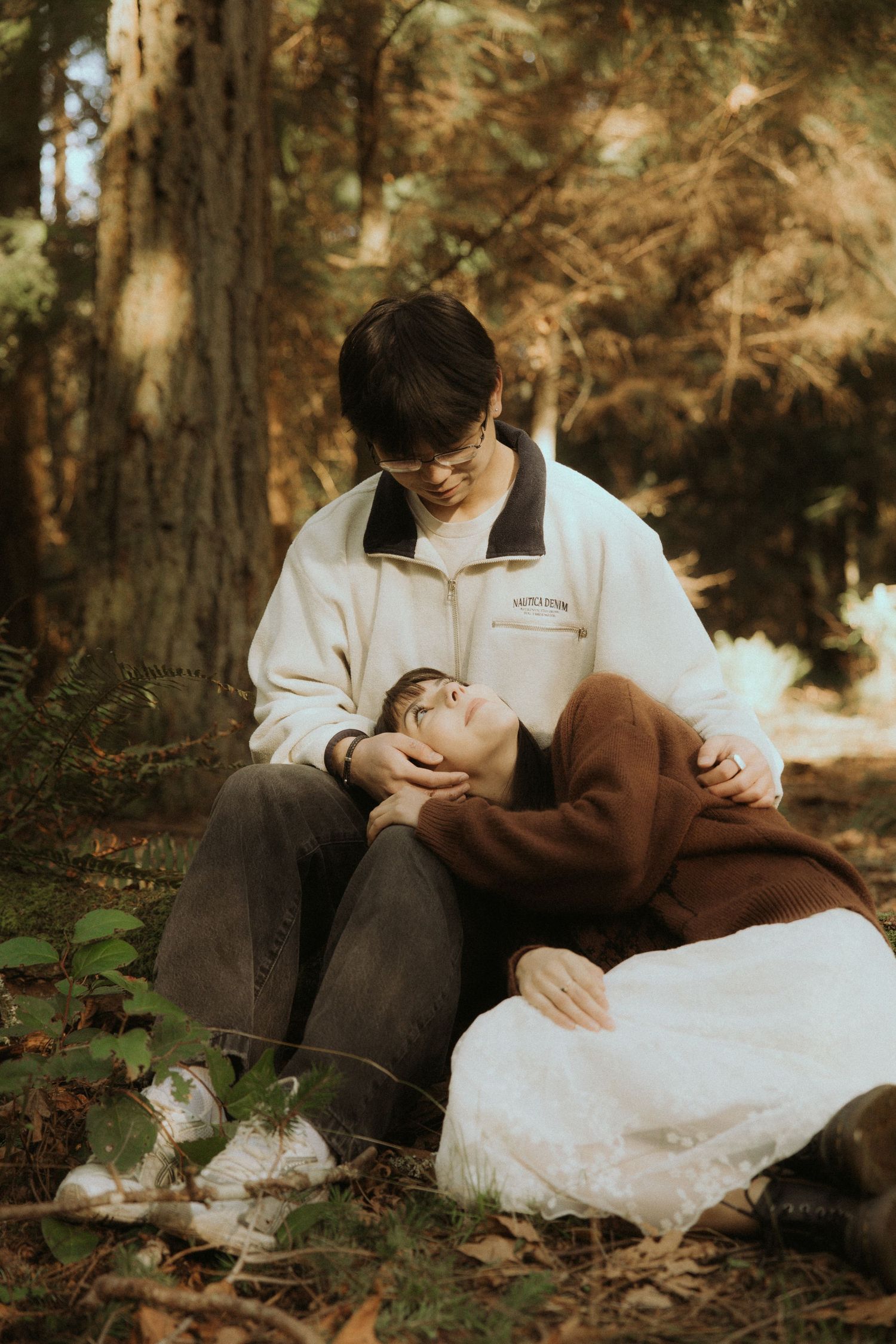A couple in brown and white attire shares an intimate moment while sitting among fallen leaves in a forest setting.