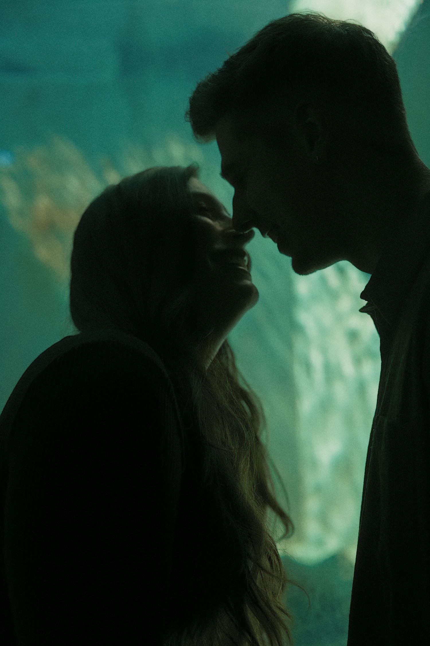 Silhouetted figures in romantic poses against a dreamy turquoise background create an intimate atmospheric scene.