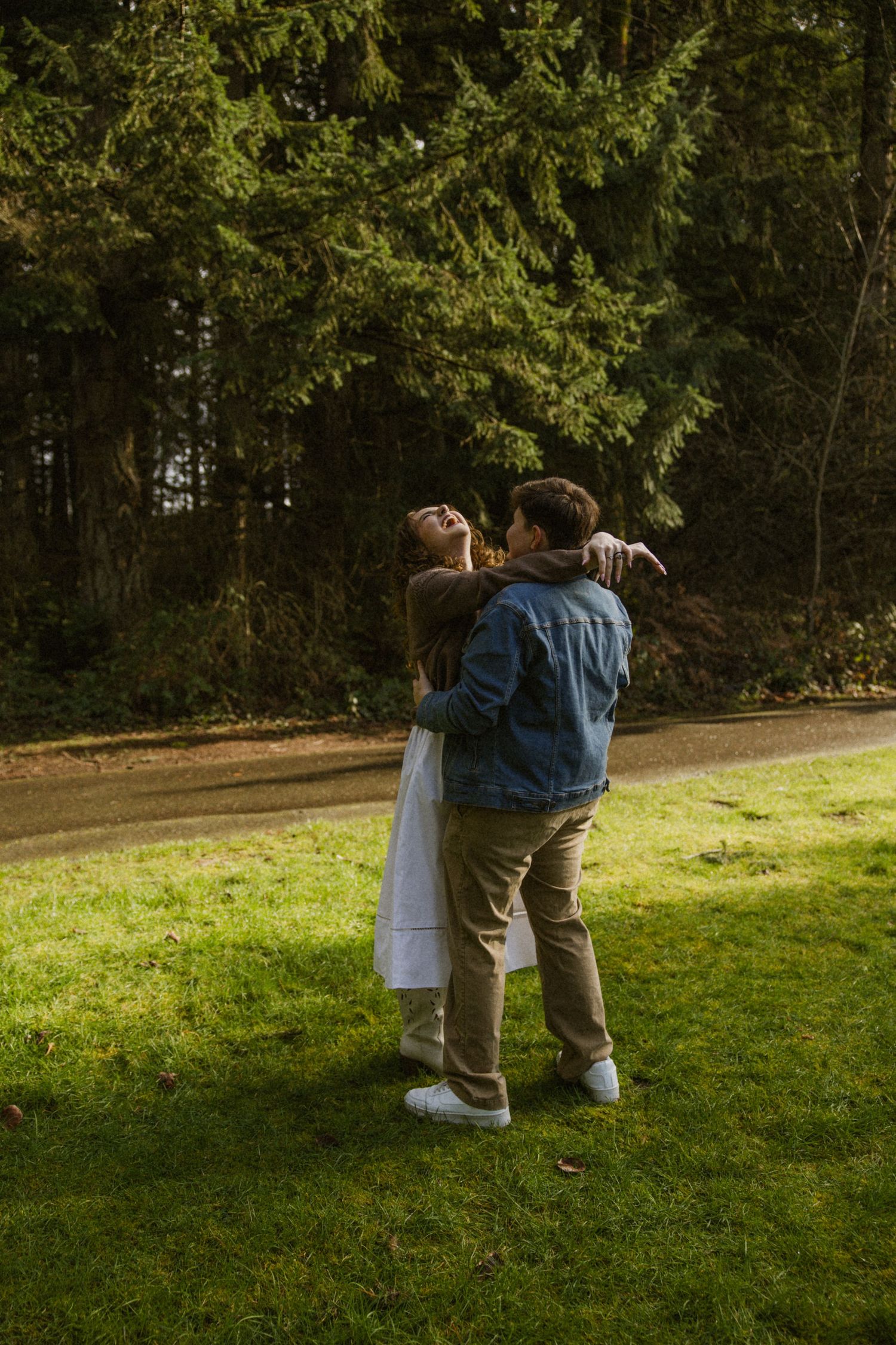A couple embraces on a grassy lawn with evergreen trees in the background during golden hour.