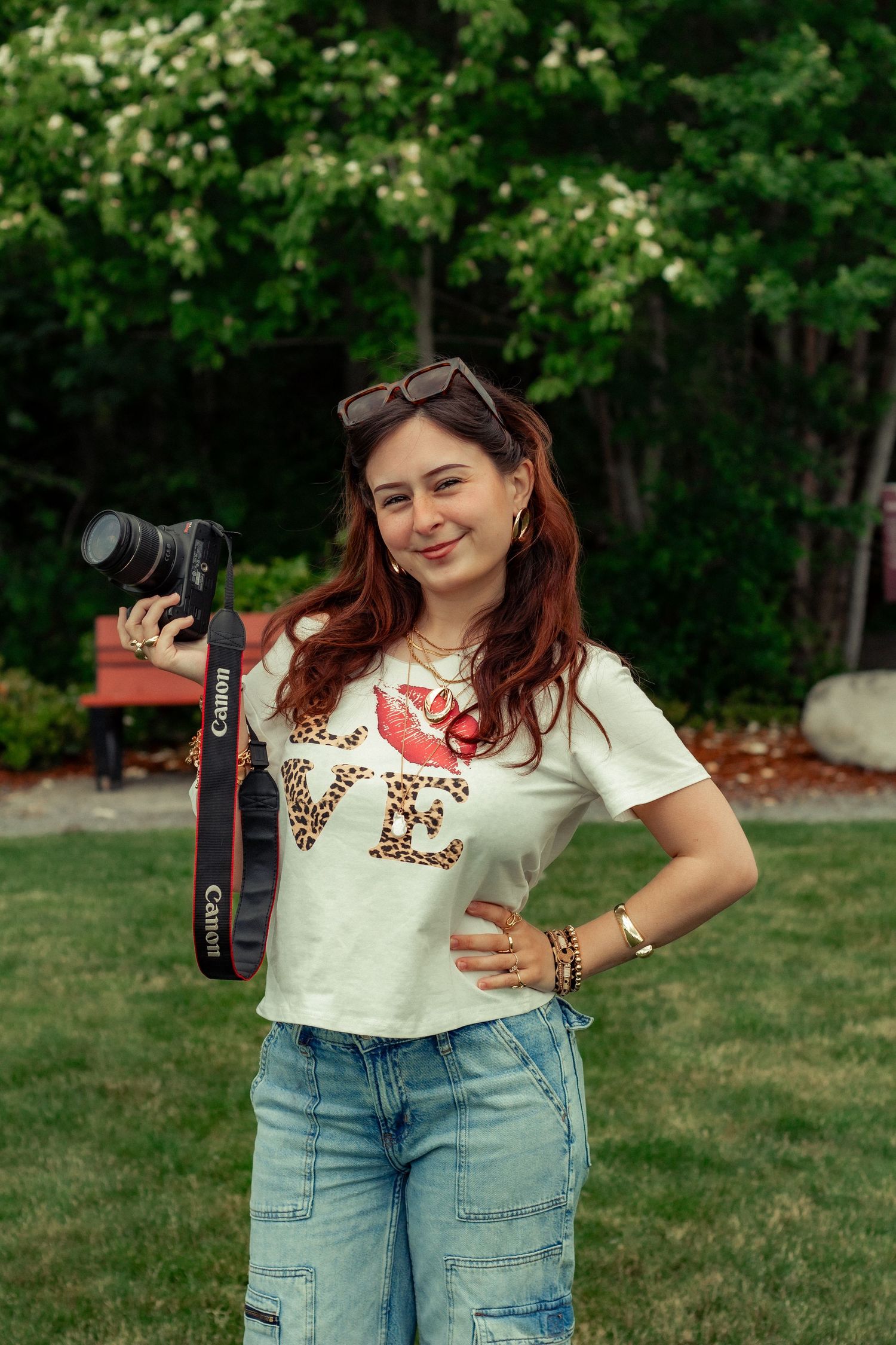 A person wearing a casual love t-shirt and jeans poses outdoors with a camera and sunglasses on their head.