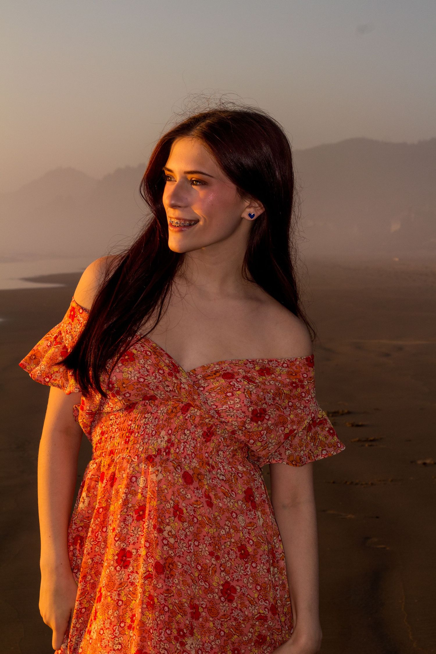 A person in a floral orange off-shoulder dress smiles while standing outdoors during golden hour.