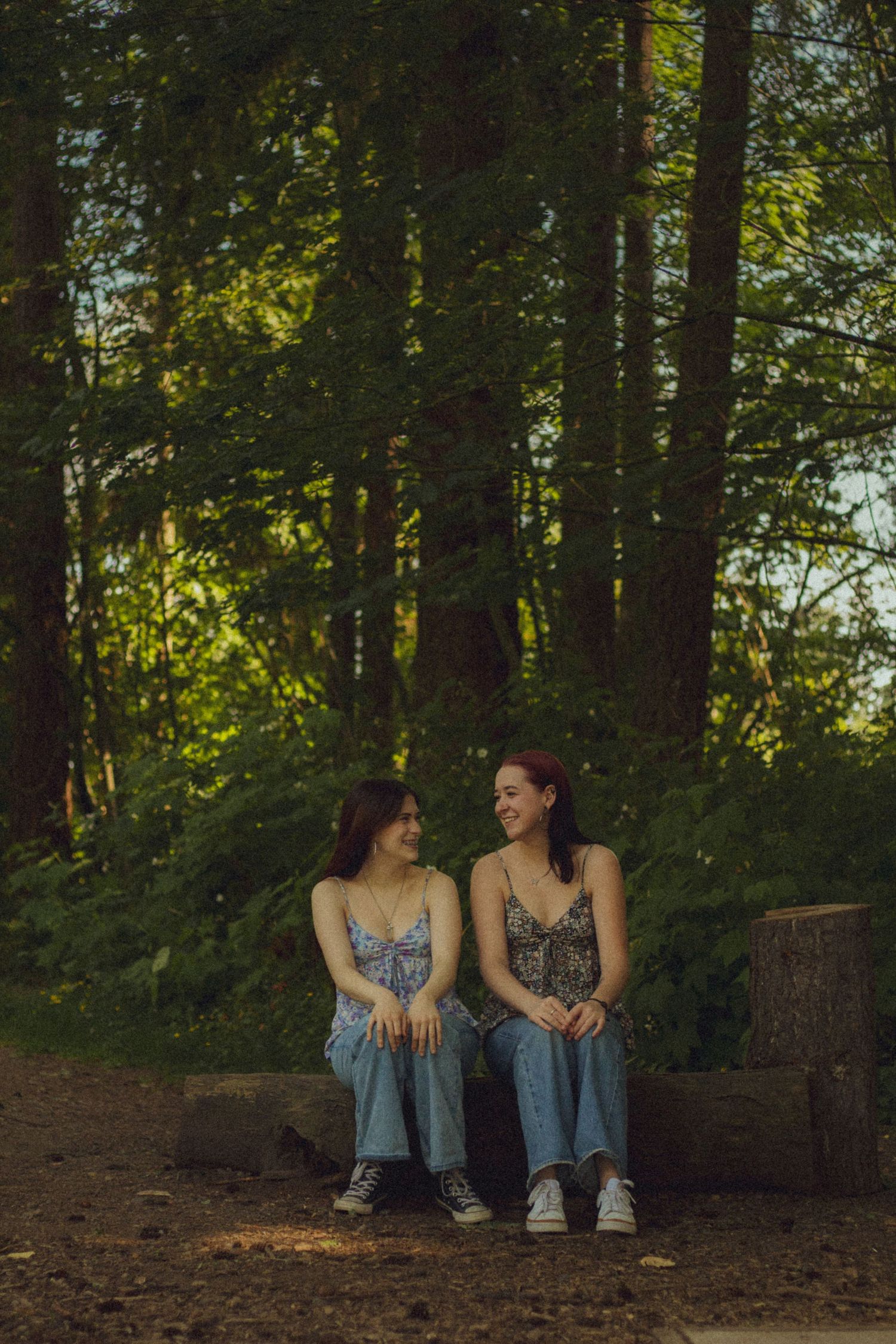 Two people in blue dresses sit together on the ground in a wooded area during golden hour.