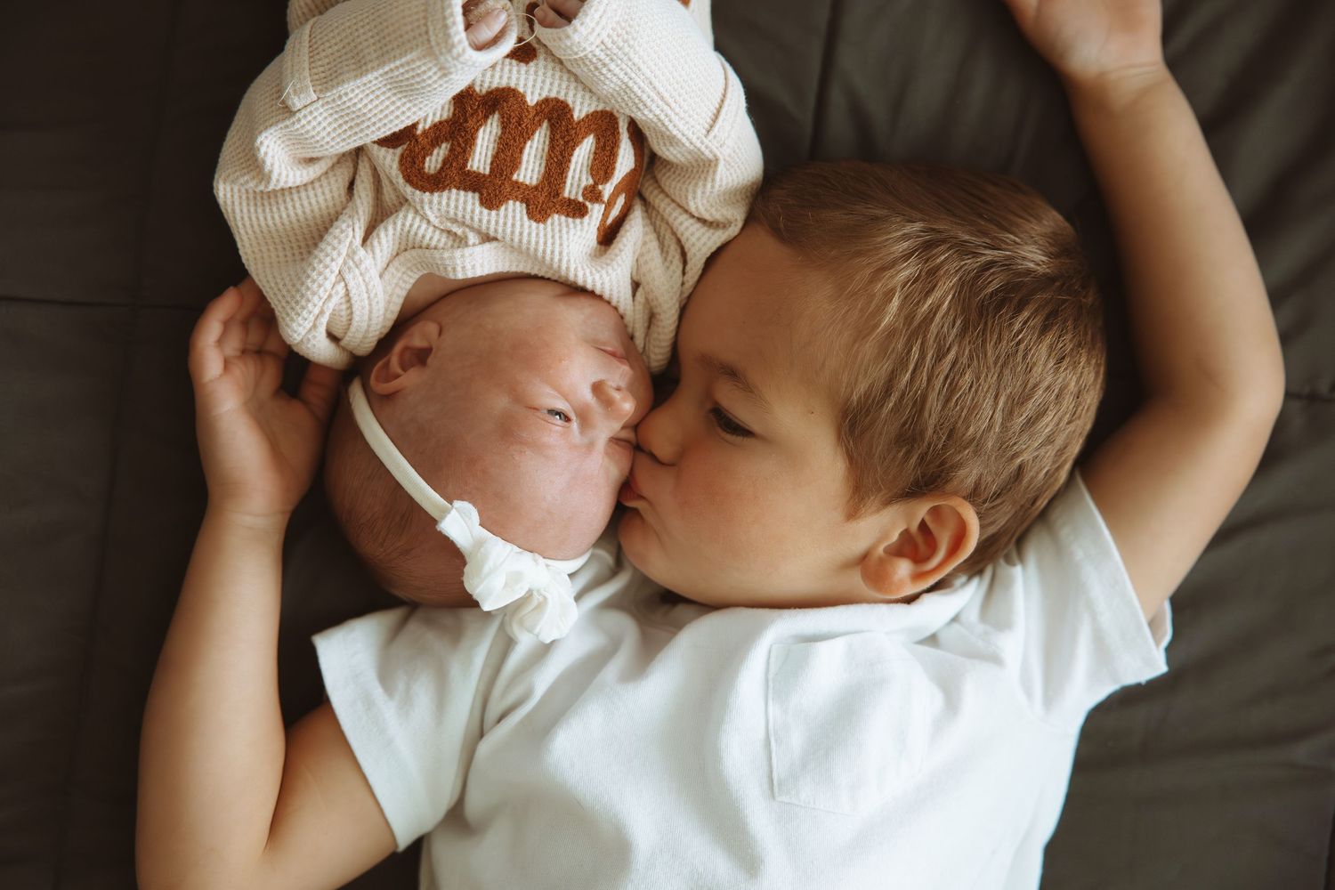 Older toddler boy in white shirt kissing newborn baby sibling wearing monogrammed hospital hat while lying together.